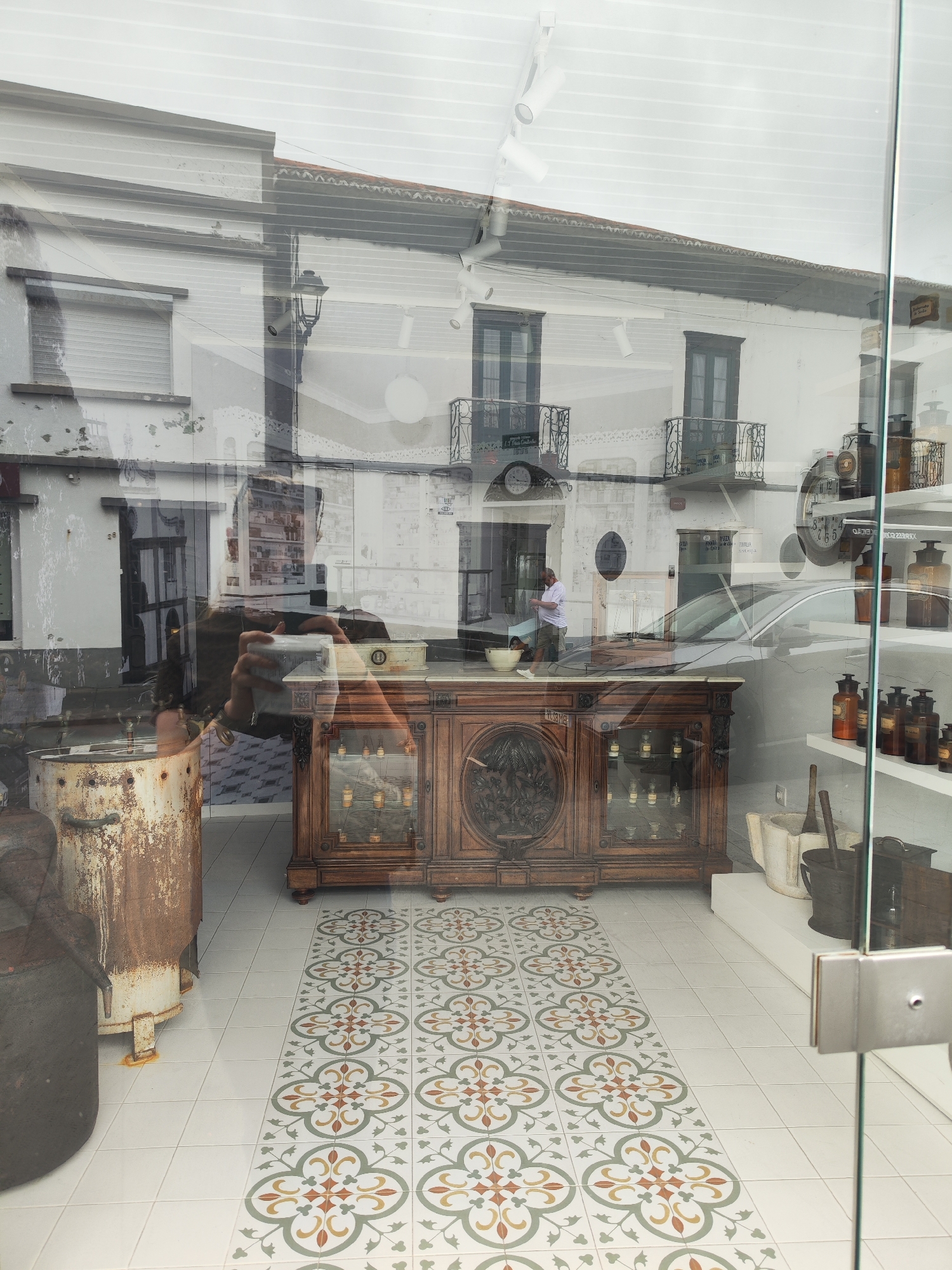 The image captures the interior of a shop in Ribeira Grande, Portugal, likely an apothecary or antique store, seen through a glass storefront. The interior features decorative tiled flooring, a large wooden cabinet with glass display cases, and shelves stocked with antique apothecary bottles and equipment. In the reflection of the glass storefront, a section of the street is visible, showing buildings with balconies, including architectural details characteristic of Ribeira Grande, and a person standing by a car. Another person is reflected holding a camera in front of the store, capturing the shop's display.