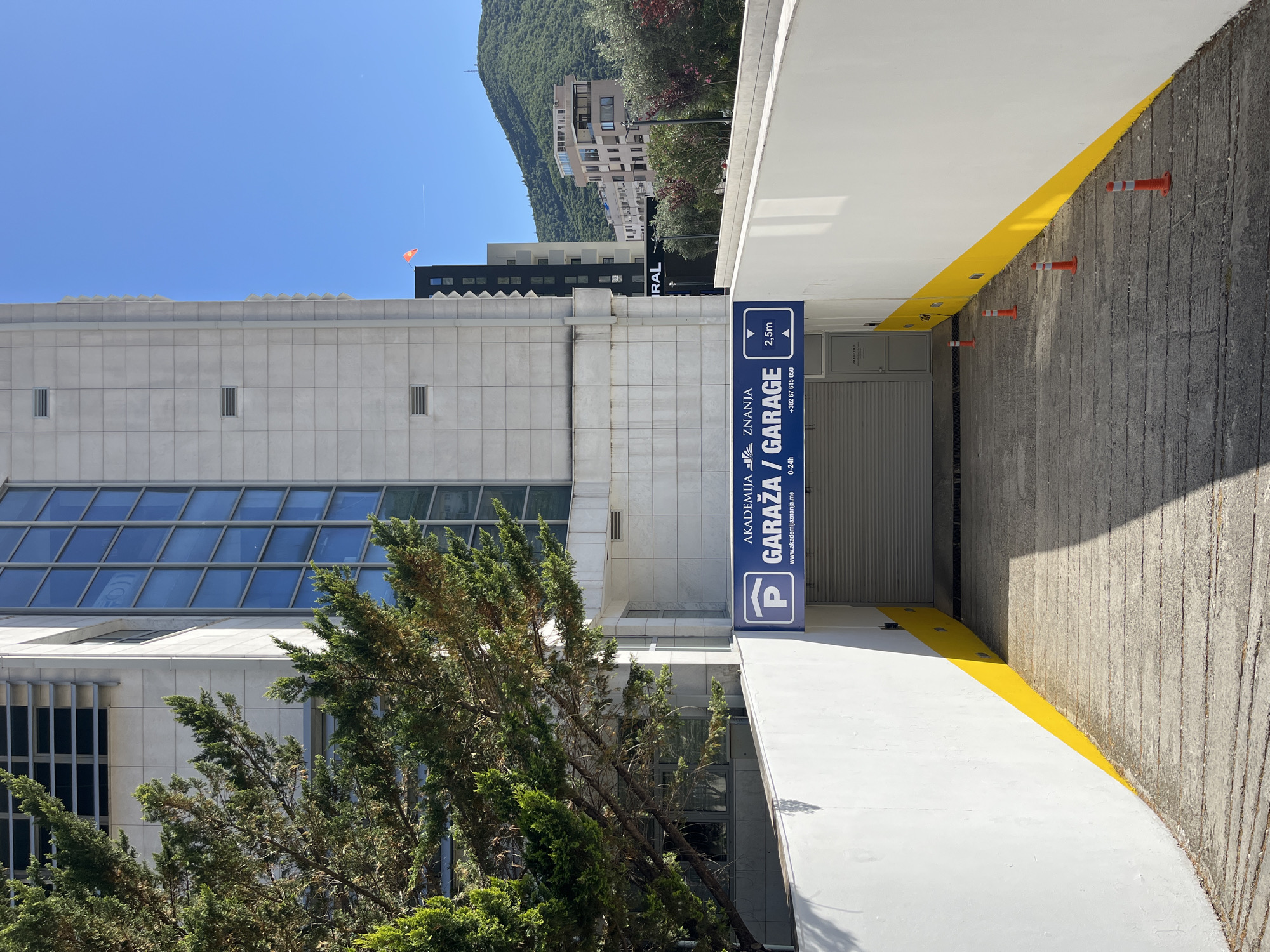 The image shows a garage entrance in Budva, Montenegro. The garage entrance has a blue sign indicating "Akademija Znanja Garage" with a parking symbol. The sign also indicates a height restriction of 2.5 meters. The entrance is flanked by white walls with a yellow stripe along the edge, with orange traffic bollards. The facade of the Academia building is visible to the left, with a glass roof and various windows. A tree with green foliage is located in front of the building. In the background, a hill with trees and buildings on it is visible under a blue sky.