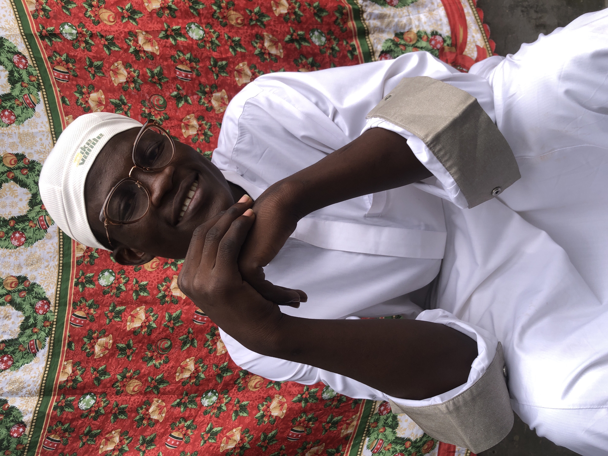 A medium shot captures a person wearing a white cap and glasses, seated and smiling in front of a red fabric with a Christmas pattern of holly, berries, bells, and ornaments. The person is dressed in a white garment with grey cuffs and has their hands clasped in front of them. The background suggests an indoor setting, possibly in Ado, Nigeria, given the festive yet casual presentation. The lighting is even, highlighting the subject and the colorful backdrop.
