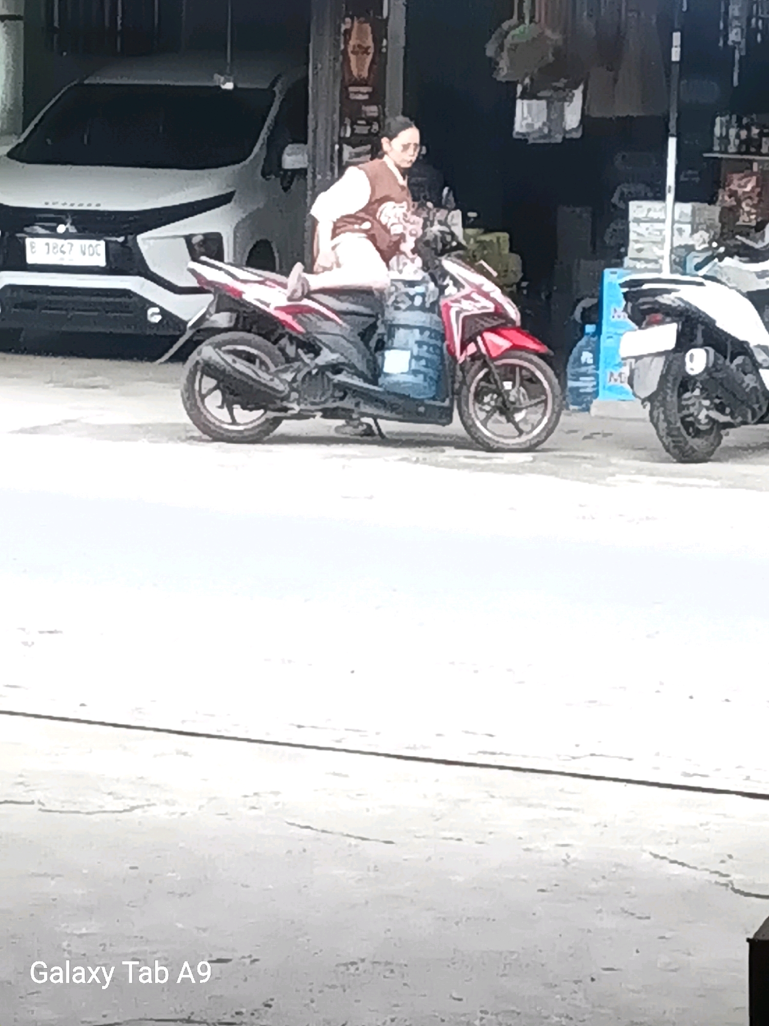 A low-angle outdoor medium shot captures a woman sitting on a red and black scooter parked in front of a store. The woman, wearing a brown vest over a light-colored shirt and shorts, is looking down and holding a bottle of water. The scooter has a large blue water jug strapped to its rear. To the left of the scooter is a white SUV with a license plate that reads "E 1867 VOC." To the right of the scooter is another scooter, partially visible. The scene appears to be in the city of Trenggalek, Indonesia, based on the visible license plate format and the presence of what looks like a typical roadside store. The ground is paved with concrete.
