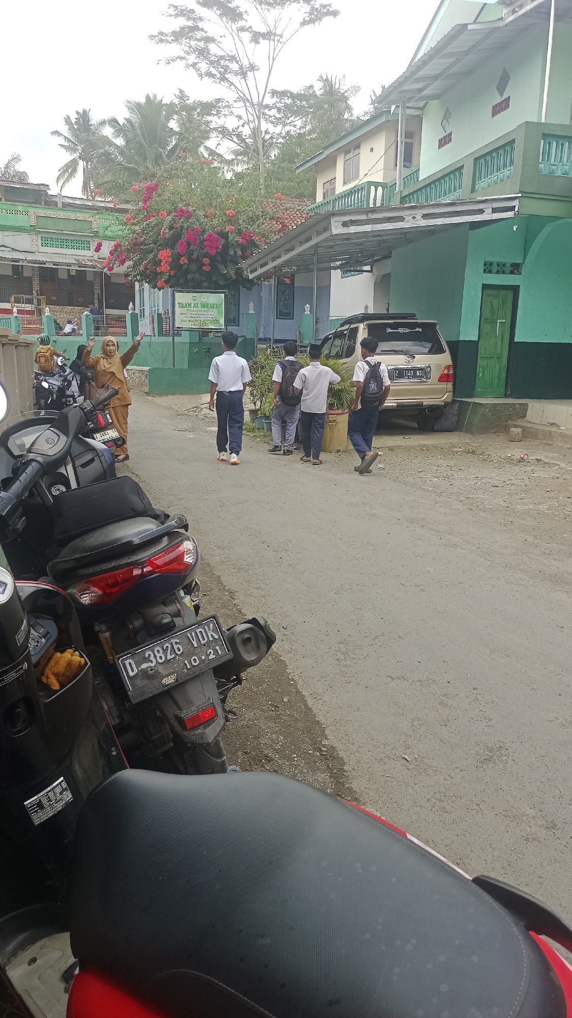 In Karyamandala, Indonesia, a dusty road in front of a light green and yellow building complex, likely a school or religious institution, is captured. Several students in white shirts and dark pants are walking away from the viewer, some with backpacks. In the foreground, motorcycles are parked. A tan van is parked on the right side of the road, near the building entrance. A woman in a tan outfit is standing to the left, gesturing with her hands raised. Colorful flowers bloom on a bush to the left of the building. The scene suggests an ordinary day at an educational facility in a rural or suburban Indonesian setting.