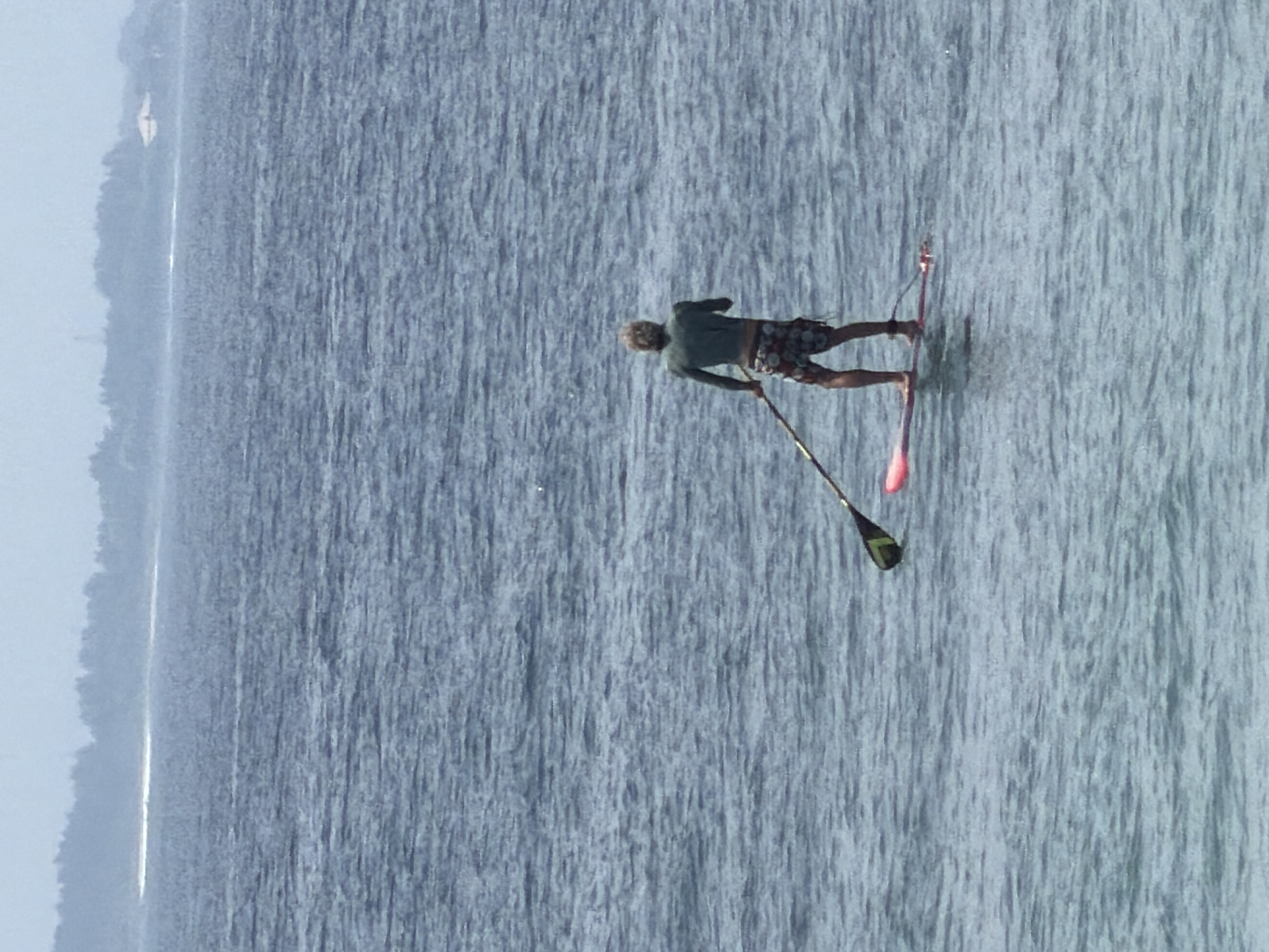 The scene depicts an individual stand-up paddleboarding in the open waters of Nusa Dua, Indonesia. An adult male, viewed from behind, is the sole human subject. He has light-colored, wavy hair and wears a dark green long-sleeved shirt and patterned swim shorts. He stands barefoot on a pink paddleboard, which is secured to his right ankle by a leash. He holds a paddle with a black and lime green blade, which is partially submerged to his left. The surrounding water exhibits a dark blue-grey hue with visible ripples across its surface. In the distant background, a hazy landmass is discernible, featuring dense vegetation, an indistinct light-colored building, and a faint white line along the horizon, possibly indicative of a distant wave break or shoreline.