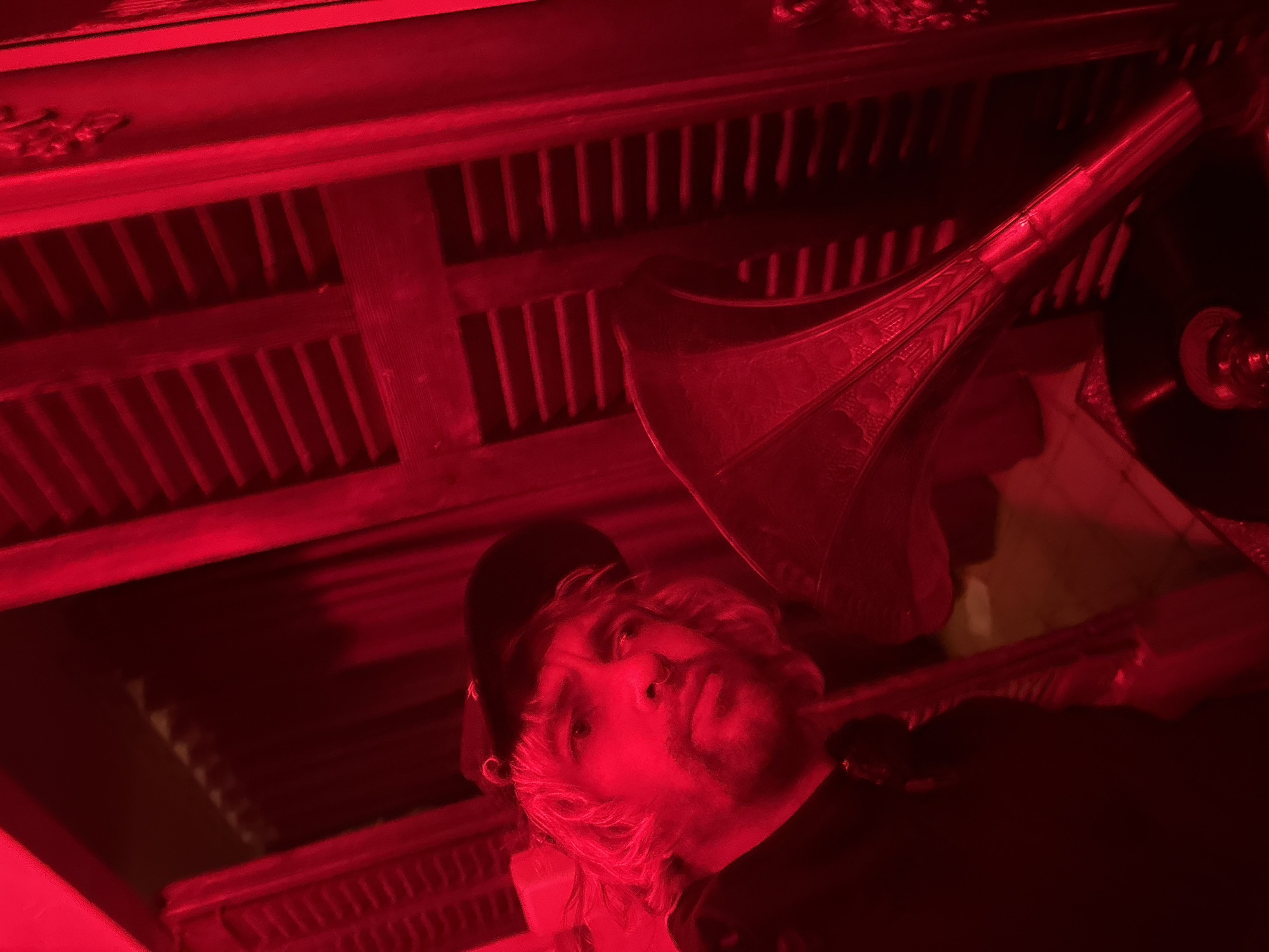 The image, bathed in a strong red light, presents a low-angle shot. It captures a person with shoulder-length, light-colored hair wearing a dark baseball cap. Positioned above and slightly behind the person is an antique-looking gramophone with a prominent horn. The background features a wooden structure with a lattice-like design. There is no information present that leads to a precise location in Tel-Aviv, Israel.