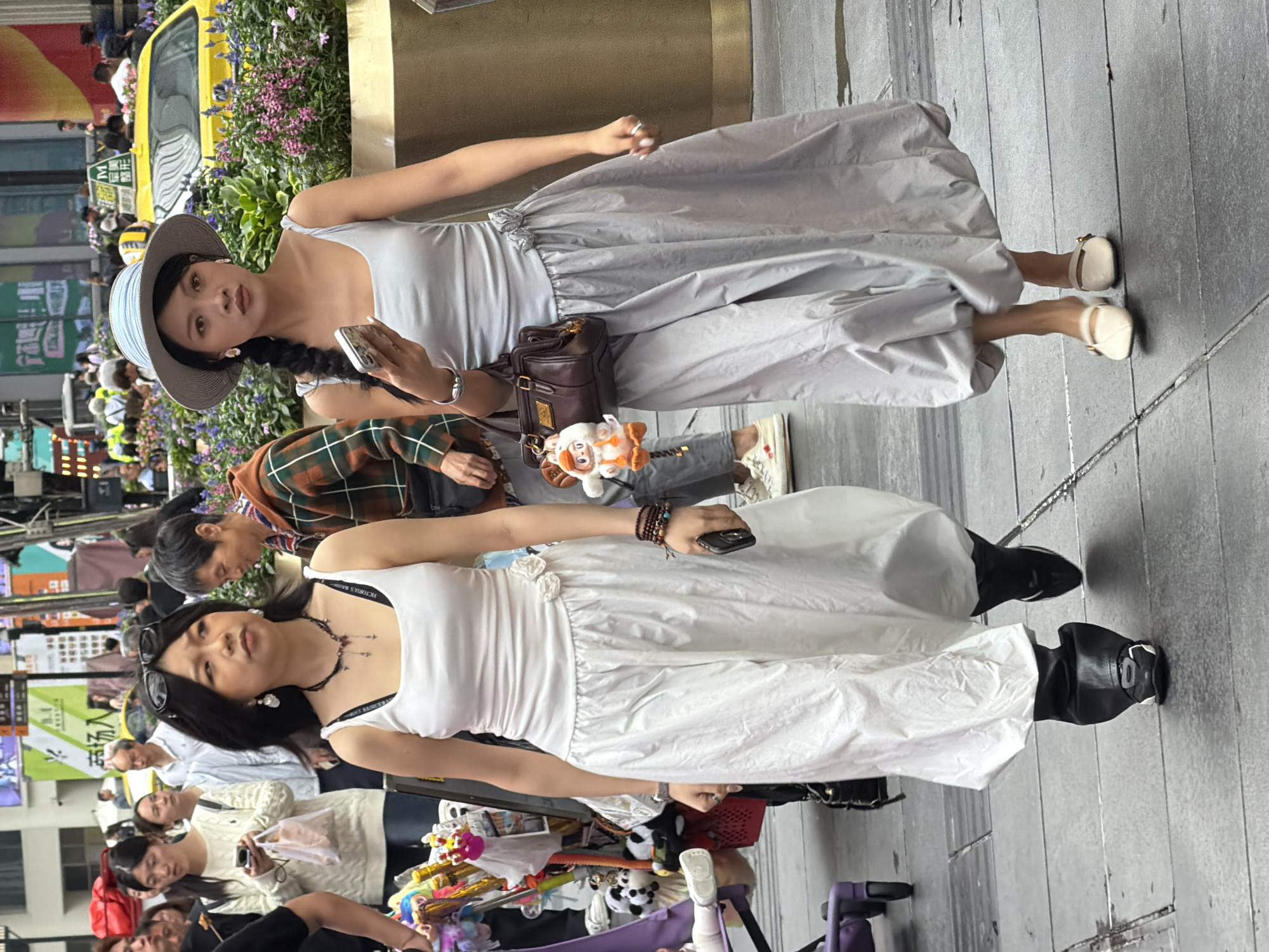 Two young Asian women walk side-by-side on a wet, paved city street in Yuzhong District, China. The woman on the left wears a white tank top and a voluminous white skirt, paired with black pants and black sneakers. She carries a dark phone in her right hand and has a dark necklace with charms. The woman on the right wears a light grey tank top and a matching grey balloon skirt, accessorized with a straw hat and white mules. She holds a cell phone in her right hand and a small brown handbag with a stuffed animal keychain hangs from her left arm. Both women appear to be looking forward. In the background, a yellow taxi is visible, along with various people and shops. The overall scene suggests a busy urban environment.