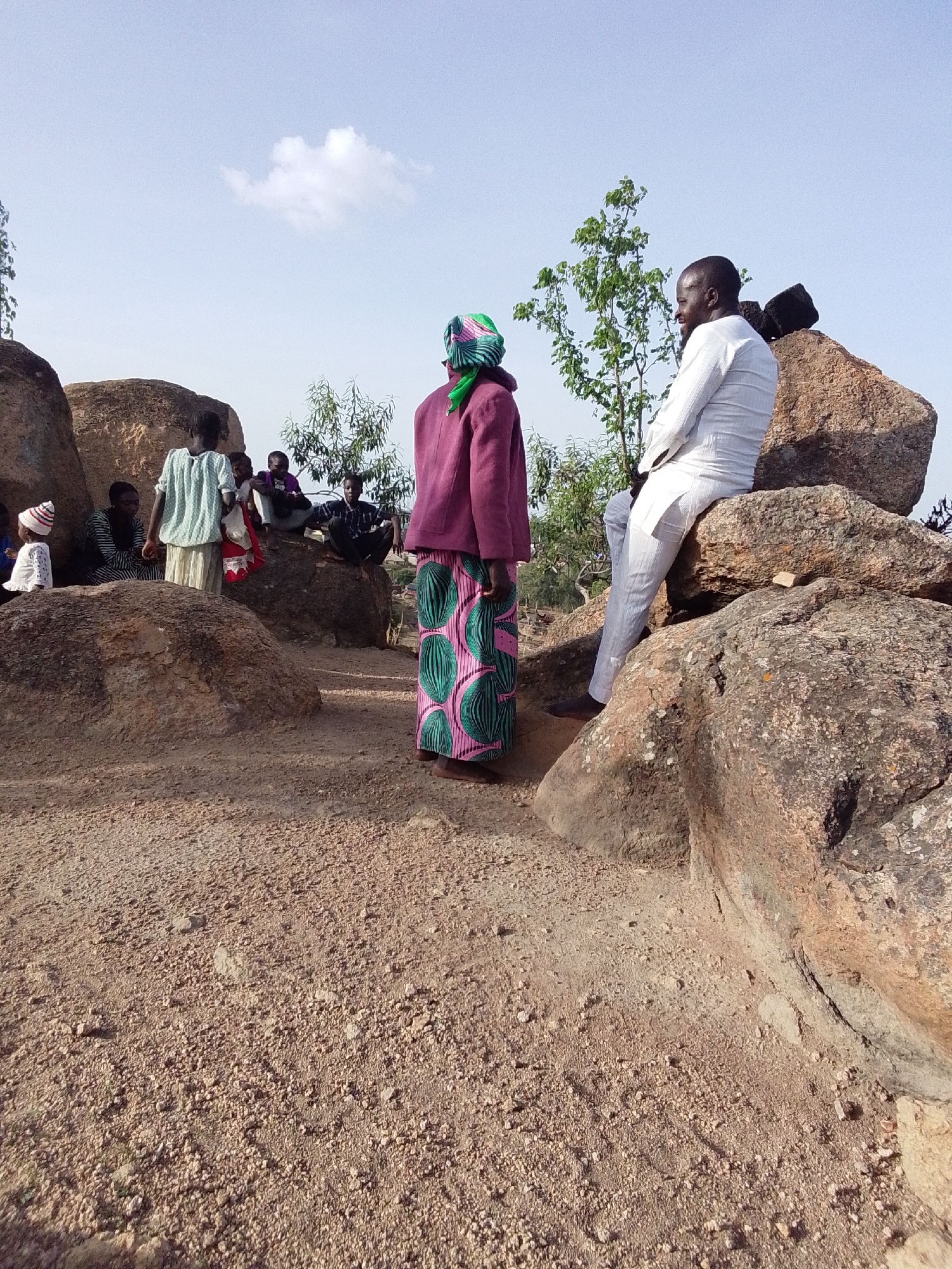 The image, taken in Barakin Akawo, Nigeria, depicts a group of people gathered on a rocky, uneven terrain under a partly cloudy sky. The ground is covered in gravel and small stones. A man in a white outfit sits on a large rock formation on the right side of the frame. He is looking to his left. A woman in a colorful skirt, a purple jacket, and a patterned headscarf stands facing away from the camera. A group of people, including women and children, are sitting or standing near another set of rocks in the background. A few small trees are also present. The scene appears to be a gathering in a natural, rural setting.