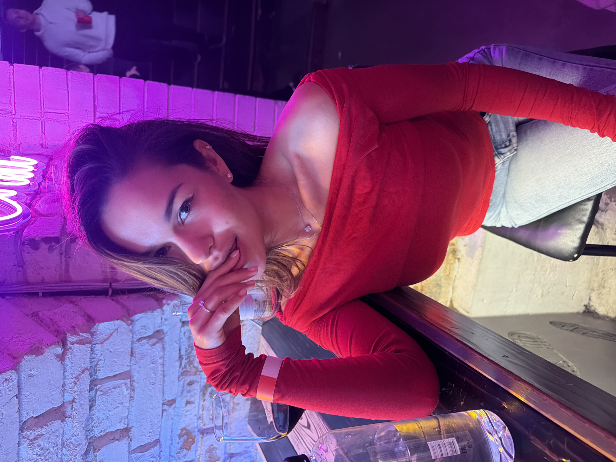 The media file displays a young woman in a vibrant, dimly lit indoor setting, likely a bar or lounge in Krasnoyarsk, Russia. The image is oriented sideways, with the woman positioned horizontally, leaning onto a dark wooden or laminate bar surface.
The main subject is a young woman with long, dark hair, styled to one side, looking directly at the camera with a slight smile and her tongue partially visible. She wears a striking red off-the-shoulder long-sleeved top and light-wash blue jeans. Her accessories include stud earrings, a delicate silver-colored necklace with a small pendant, and a ring on her right index finger. An orange and white wristband is visible on her left wrist.
On the bar surface in front of her are an empty clear drinking glass and a plastic water bottle. The bottle clearly shows partial text including "0.5L", "EAN 380...", and "0.2..." along with a barcode. The background features a brick wall, partially painted white and fading into a muted pink/purple hue, strongly illuminated by a purple neon light. Part of a neon sign, possibly spelling "LOUNGE" or a similar word, is visible but cropped. Above the wall, a blurry reflection of a person in a white hoodie is discernible, suggesting a mirrored surface or distant glass. The overall lighting is artificial and predominantly purple, casting a cool, moody ambiance. The woman appears relaxed and confident, posing for the photograph in what seems to be an evening or nighttime environment.