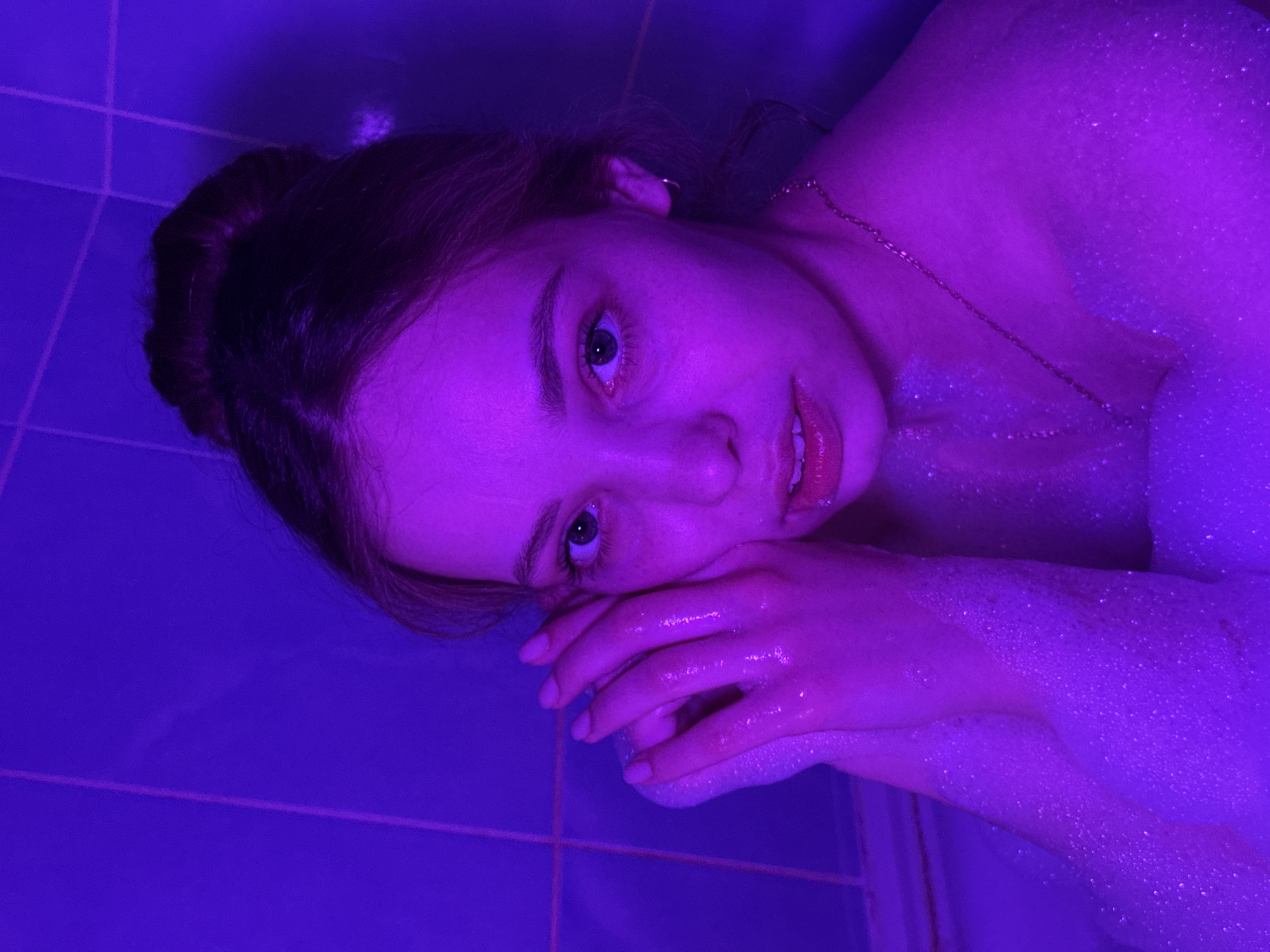 A woman is lying in a bathtub, partially submerged in soapy water. She is fair-skinned with light eyes and dark hair pulled back in a bun. She is wearing a delicate gold necklace. Her hands are resting on the edge of the tub. The scene is illuminated with an intense purple light. The bathtub and the surrounding tiling are also purple. The location is the city of Chaikovsky, Russia.