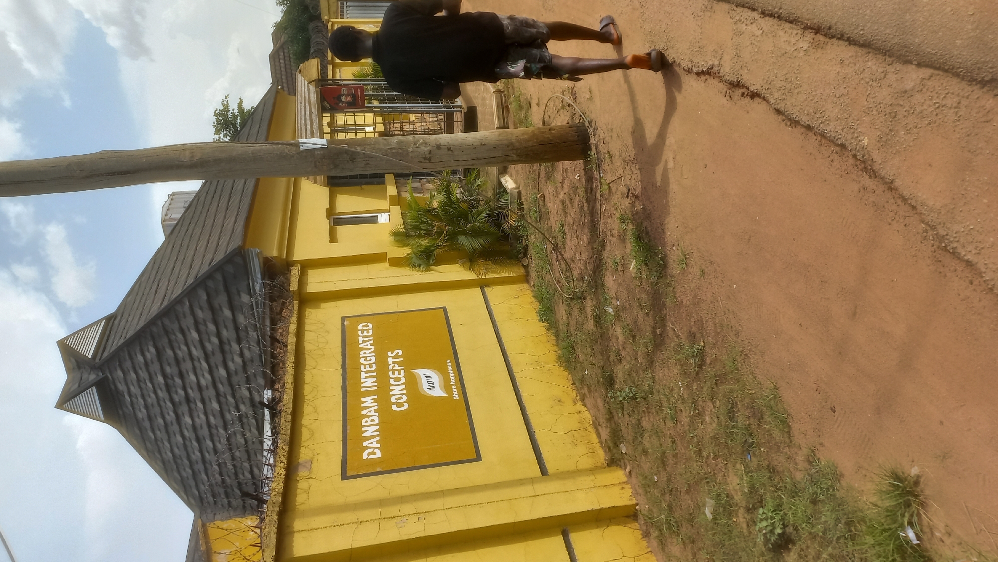 The image depicts the exterior of a building with a distinctive pyramidal roof, painted bright yellow. A sign on the wall reads "DANBAM INTEGRATED CONCEPTS" with a logo and the tagline "Share happiness." The building is enclosed by a yellow wall topped with barbed wire. To the right of the building, a person wearing a black t-shirt and patterned shorts walks away from the camera on a dirt path. A wooden utility pole stands prominently in the foreground. The environment is outdoors, with dry grass and dirt surrounding the path and building. The sky above is partly cloudy. This scene is located in Gura Top, Nigeria.