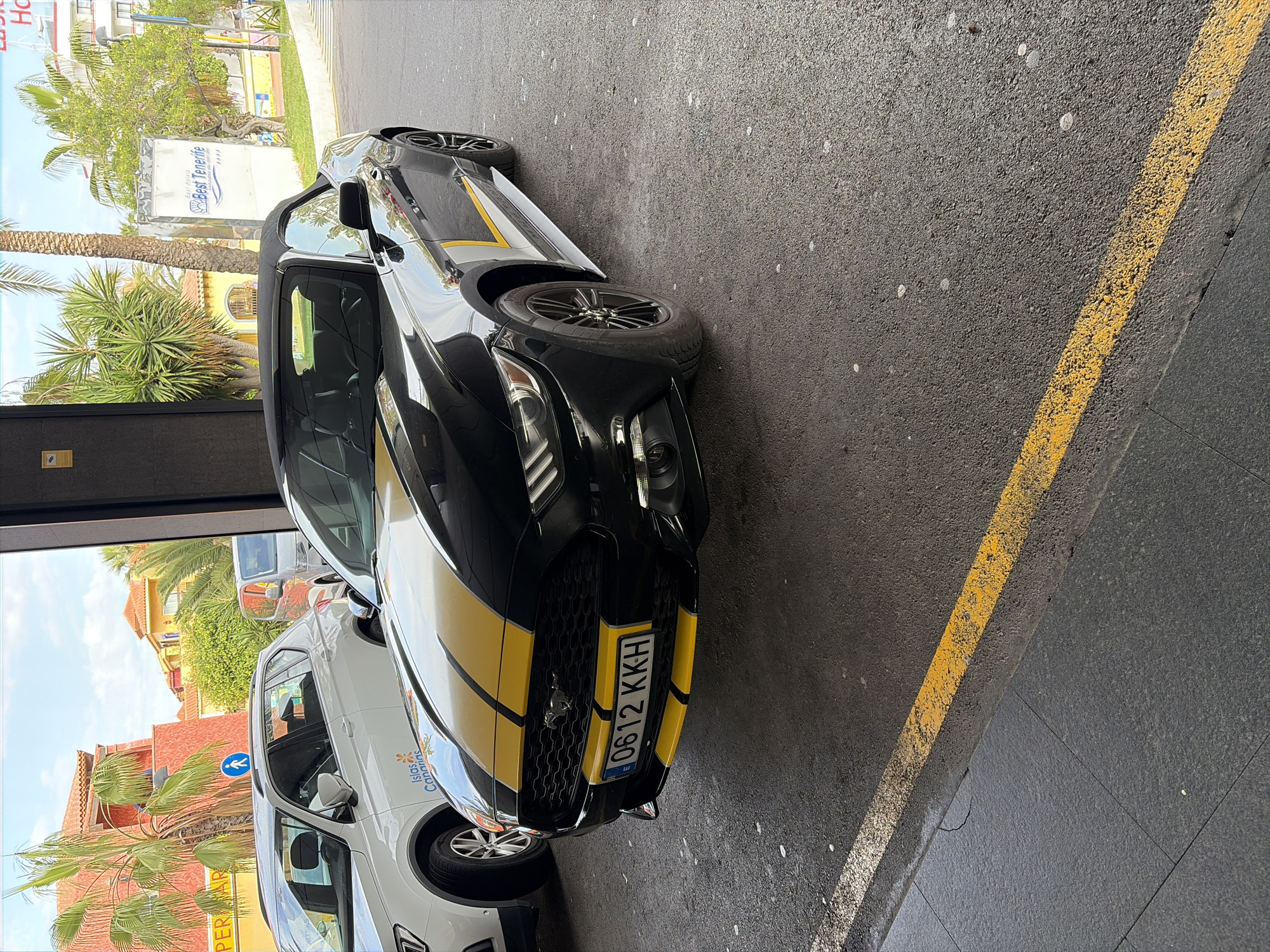 A black convertible car with yellow stripes is parked in what appears to be a parking area. A white car with the logo of "Islas Canarias" is parked beside it. The license plate of the convertible is "0612 KKH." Outside the parking area, palm trees are visible, along with buildings and a sign that reads "Best Tenerife." The scene likely takes place in Los Cristianos, Spain, given the presence of the Best Tenerife hotel, which has a branch in that city. A yellow line is painted on the road adjacent to the parking space. The image appears to be taken from inside a building looking outwards.