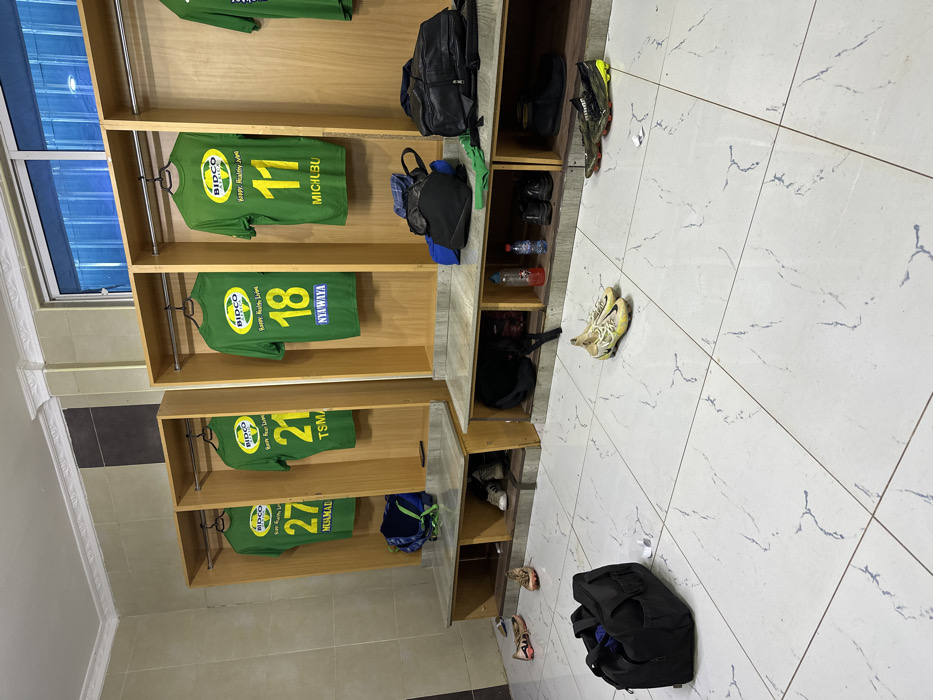 The image shows a locker room at a sports facility in Machakos, Kenya, located at coordinates -1.526 latitude and 37.26 longitude. The locker room has white tile flooring and three rows of lockers along the wall. Each locker is made of wood and has a shelf. The lockers are numbered and have green and yellow soccer jerseys hanging on them. The jerseys all have the same logo: a logo with the words "BIDCO" and "Happy, Healthy Lives" on it. The lockers are numbered 27, 21, 18 and 11. The numbers are printed in black and white on the front of each jersey. Two black duffel bags and a pair of black sneakers are on the top of the lockers. A green and yellow towel is lying on the top of one of the lockers. There are also two pairs of soccer cleats and several other pairs of shoes on the floor of the locker room.
