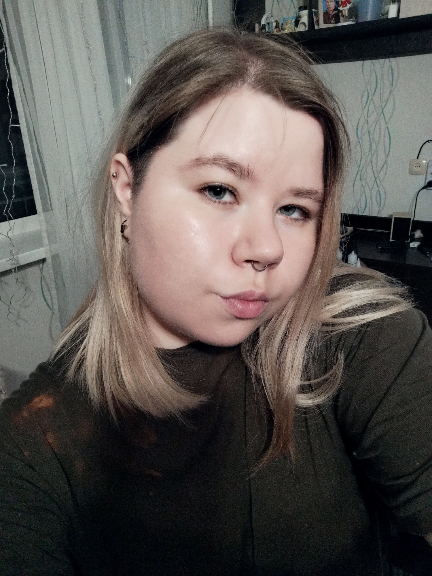 A selfie depicts a fair-skinned individual with blonde hair, blue eyes, and a nose ring. The person is wearing a dark green shirt. A shelf with various items is visible in the background. A window with white curtains is to the left. The location is Unknown, Unknown.