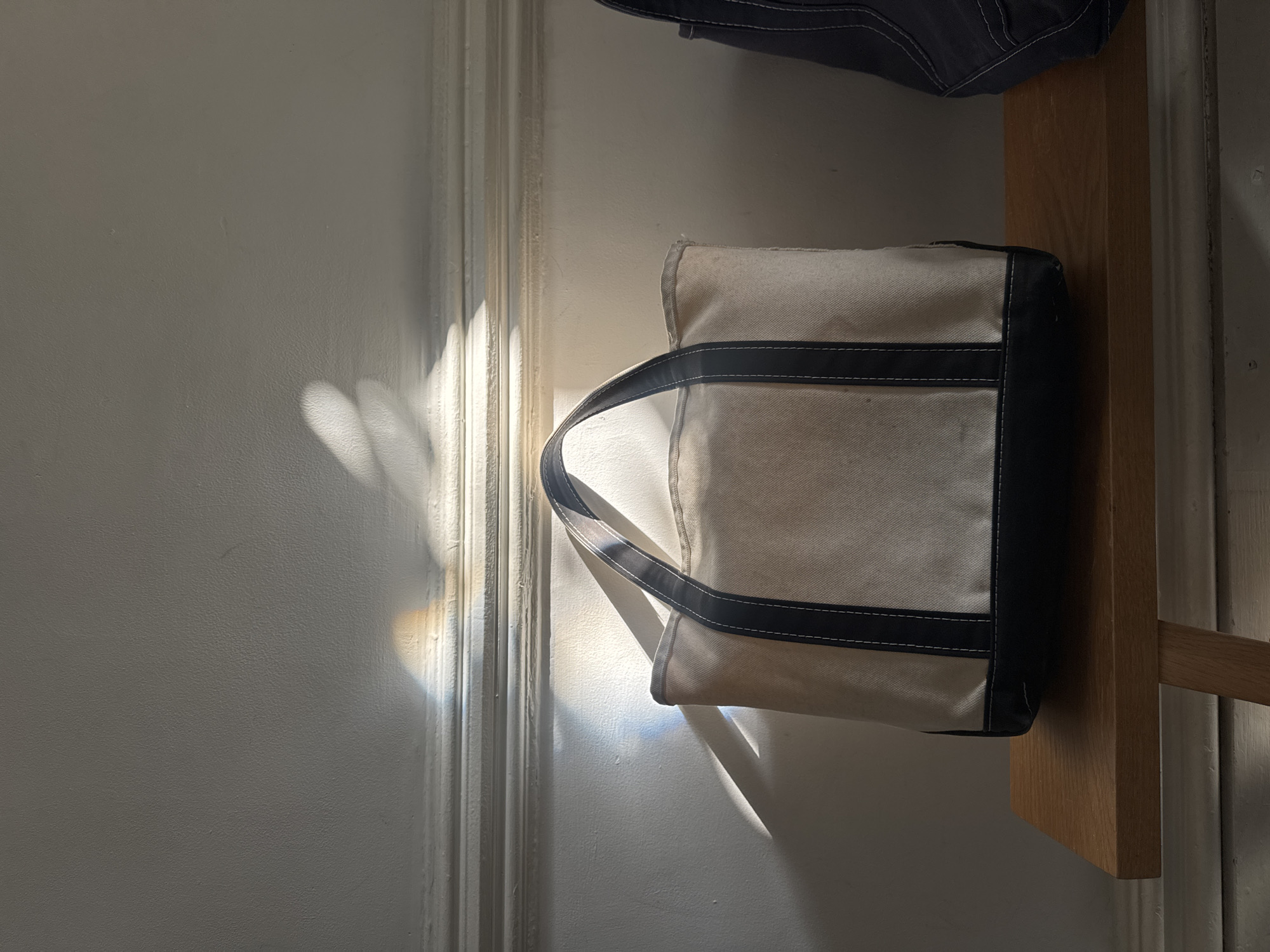 A top-down, interior shot reveals a beige canvas tote bag with navy blue trim and handles, hanging on a wooden shelf. The bag is mostly visible, situated in the upper right-hand quadrant of the frame. Parts of another dark blue object are faintly visible at the top of the frame. The background is a pale gray wall with vertical molding to the left, catching the sunlight in such a way that it creates bright patterns on the surface. The scene is bathed in natural light, giving a high-key feel to the image. Given the neutral tones and simple composition, it is difficult to identify the specific location within New York City, United States of America.