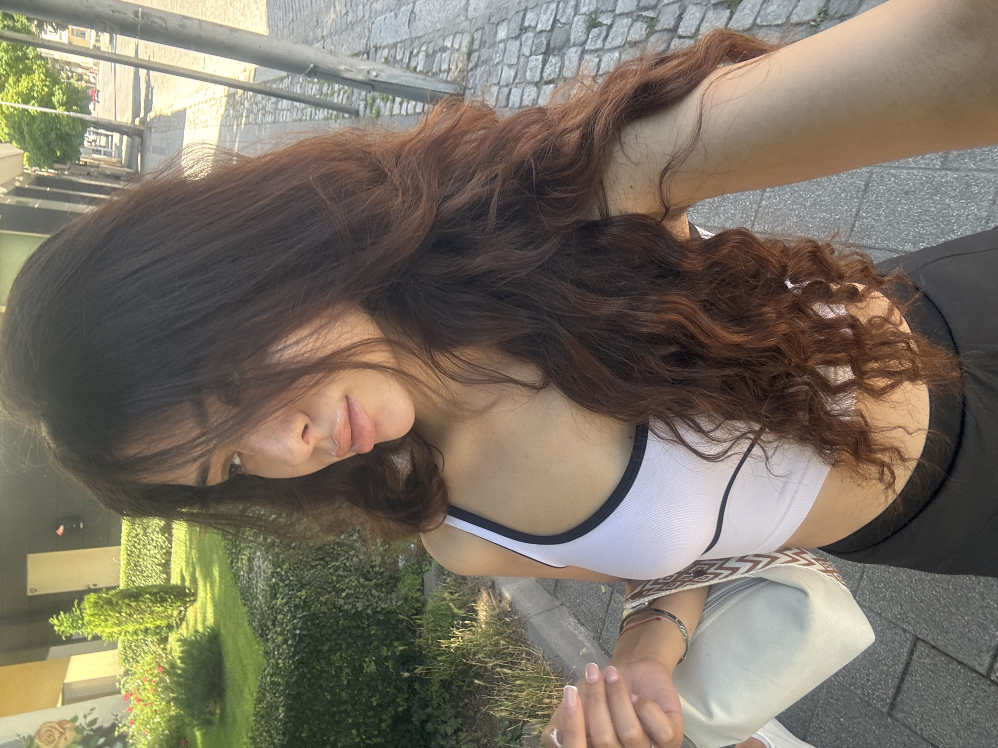 A young woman with long, wavy, dark brown hair is standing on a cobblestone sidewalk, looking to her left. She is wearing a white cropped tank top with black trim and black bottoms. She is carrying a light-colored shoulder bag. In the background, there is a well-maintained garden with green bushes and red flowers, a building with a yellow facade, and trees lining the street. The scene appears to be a sunny day on a street in Plovdiv, Bulgaria.