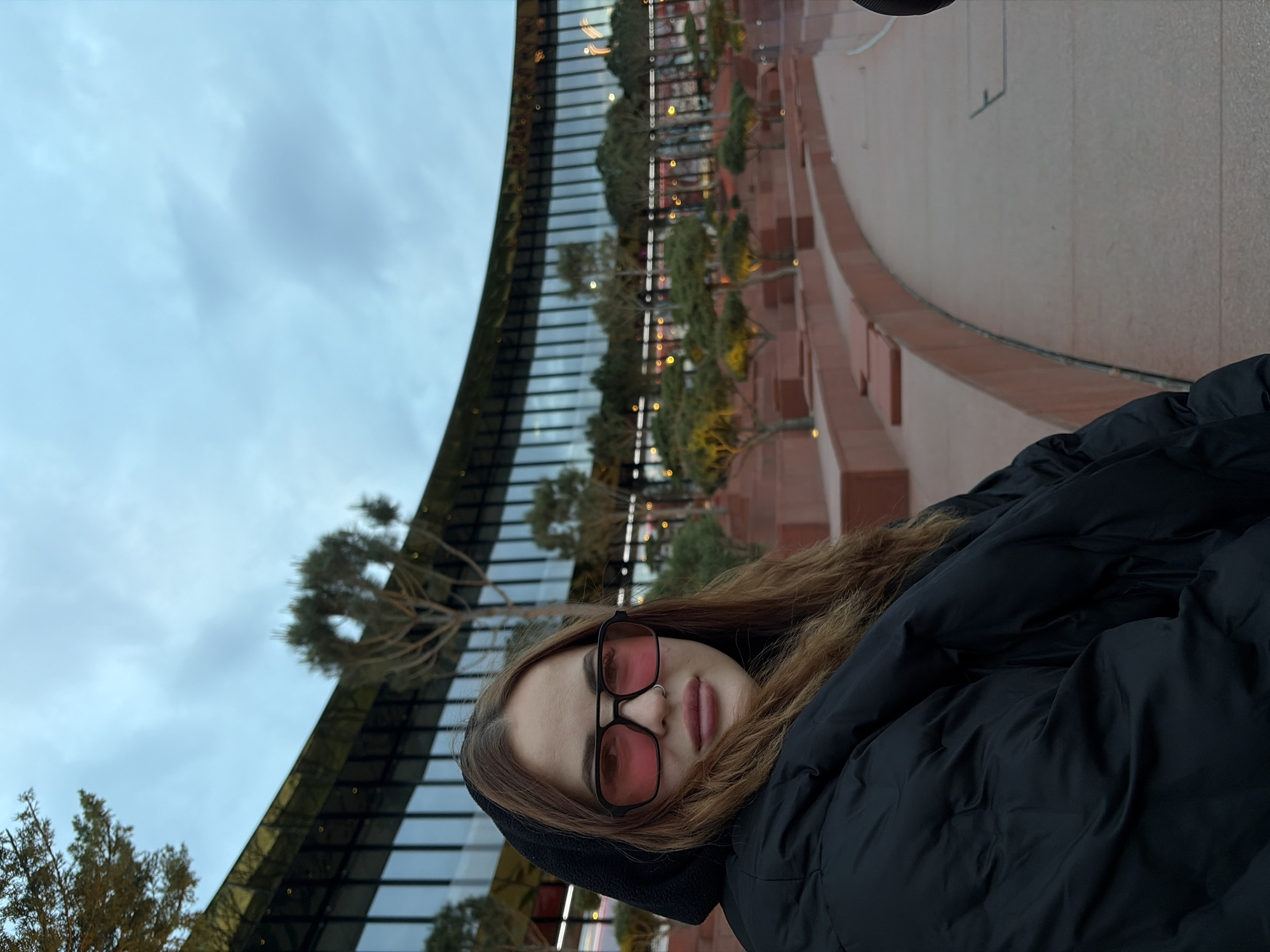 A photograph shows a woman with long brown hair, wearing black framed glasses with red tinted lenses, a black hooded jacket and a nose ring. The woman is standing on a balcony or rooftop of a building. The photo is taken outdoors, with a view of the cityscape in the background. In the background is a large building with a curved design. The building is made of glass and metal, and it has a row of trees planted on the roof. The sky is cloudy and the light is soft. The image is taken at an angle. The location is Unknown, Unknown.