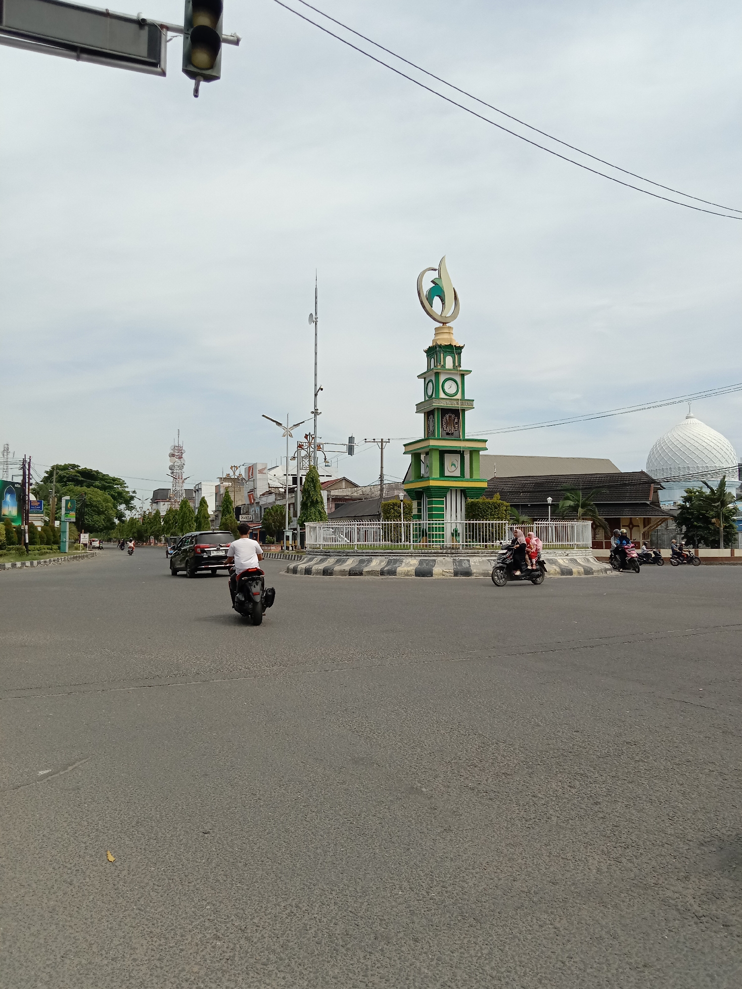 The image depicts a street scene in Lhokseumawe, Indonesia. In the center is a roundabout with a clock tower structure painted in green and yellow. The tower is topped with a religious symbol. A white mosque dome is visible behind the roundabout. Several motorbikes are on the road, some circling the roundabout. A black car is also on the road. A traffic light is visible in the upper left corner of the frame. The sky is overcast. The road surface is asphalt. Buildings and trees are visible in the background.