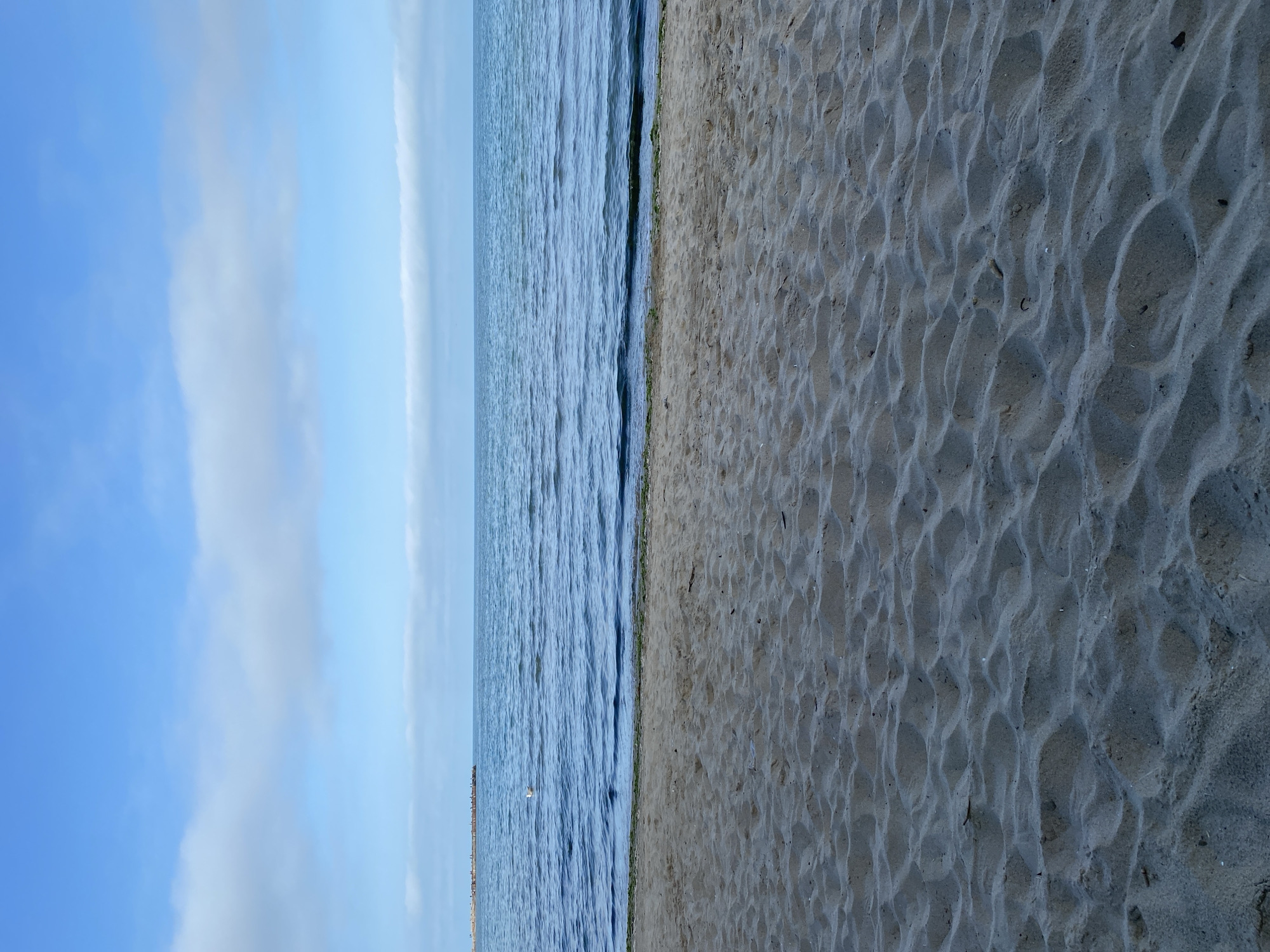 The image depicts a beach scene in Varna, Bulgaria. The composition is divided vertically, with the left side showing a blue sky with scattered clouds and the sea, while the right side displays a sandy beach. The sea has small waves, and a distant pier is faintly visible. The beach sand is textured with numerous small indentations. There are no people visible in the scene.