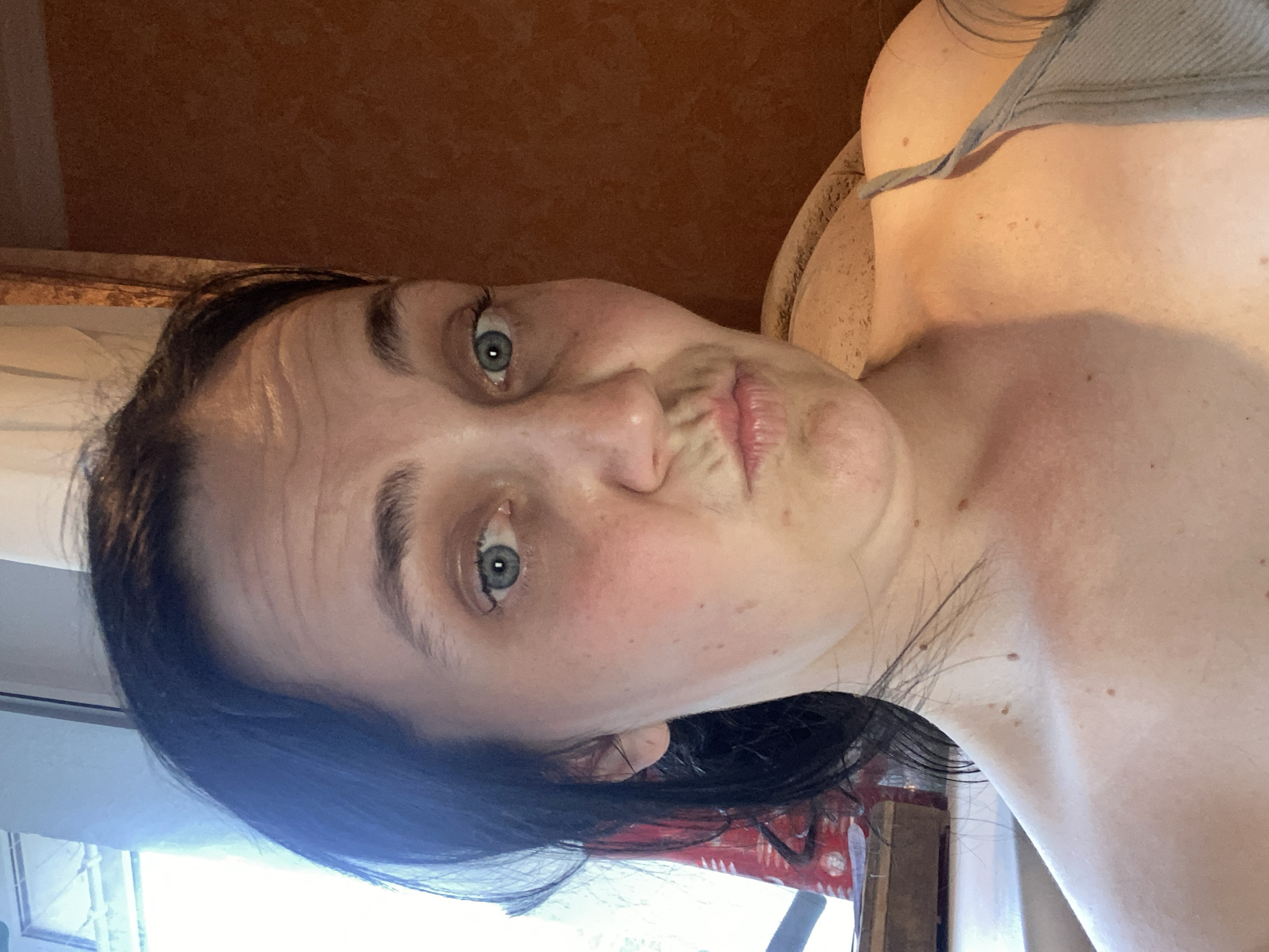 A medium shot captures a fair-skinned individual with dark hair, making a duck face for a selfie. The person's eyes are a striking blue, and their skin shows some freckles and redness. Their mouth is pursed, and horizontal lines are visible across their forehead. The background features a window with light filtering through on the left and a textured, brown wall on the right. The person is wearing a gray tank top, and part of their shoulder and chest are visible, adorned with a few small moles. The setting appears to be indoors, with the quality of light suggesting daytime. The individual is positioned in the foreground, with the background slightly out of focus.