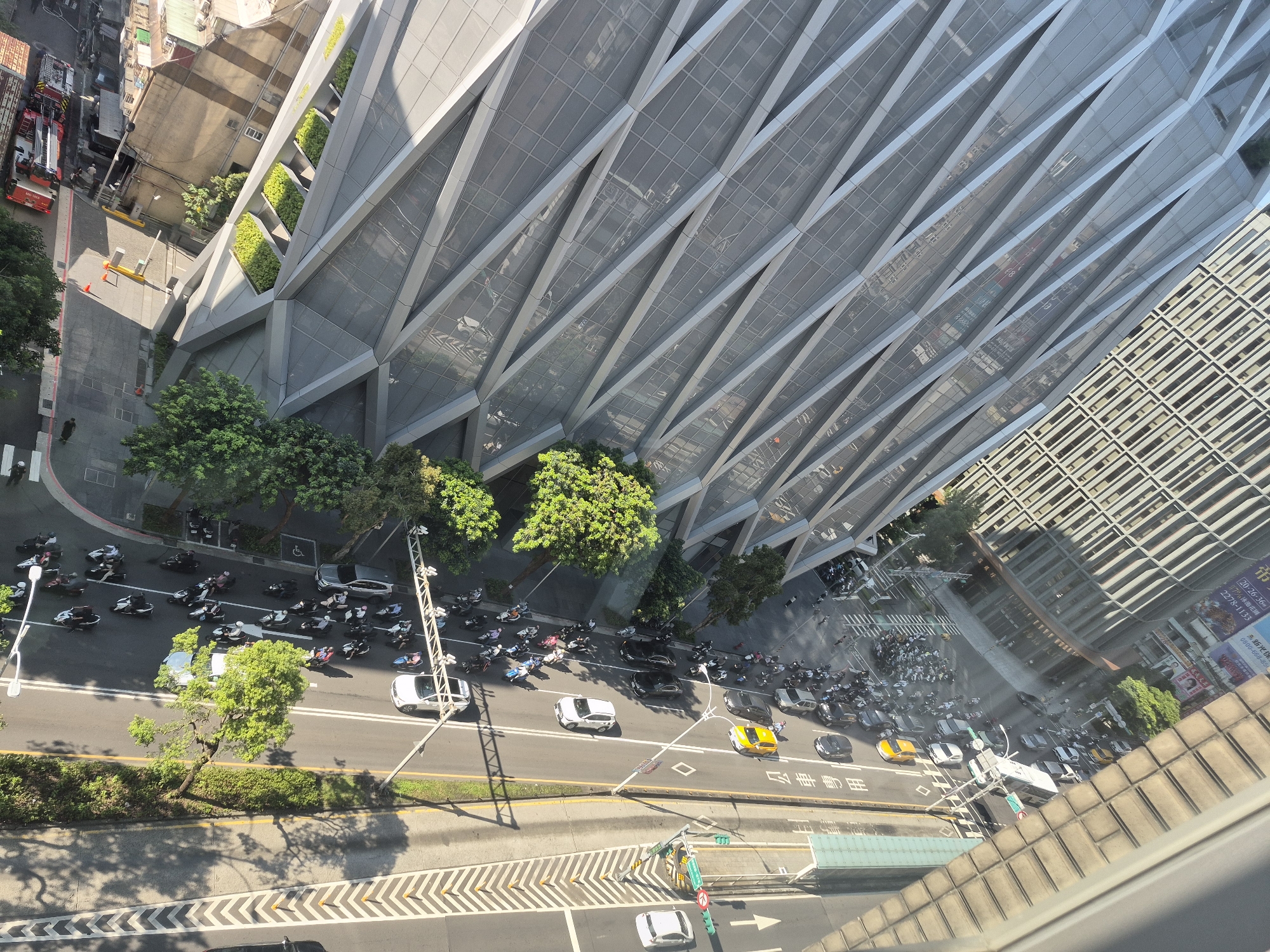 This is a high-angle aerial view of a street scene in Taipei, Taiwan. A modern, angular building with reflective glass panels dominates the right side of the frame, casting shadows on the street below. To the left of this building, other structures are visible, including an older building with a patterned facade and a red fire truck parked on the street.
The street itself is busy with traffic. Numerous motorcycles are parked on the side of the road, and many more are in motion, navigating the lanes. Several cars are also present, including a white sedan, a yellow sedan, and a dark SUV. A white car is parked in the middle of an intersection.
Trees line the street, providing shade and greenery. The environment appears to be an urban setting, with typical city infrastructure such as traffic lights and street signs. The overall impression is one of a dynamic and bustling urban environment in Taipei.