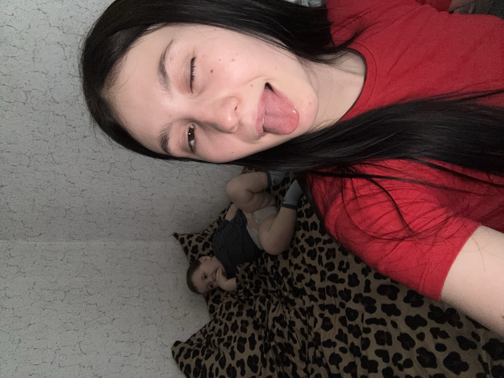 An indoor photograph captures a playful moment between a young woman and a baby. In the foreground, a woman with long dark hair, wearing a red short-sleeved top, is taking a selfie. She makes a humorous expression, winking with her right eye and sticking out her tongue. Her face shows some light freckles.
In the background, positioned slightly below and behind the woman, a baby or toddler lies on a brown and black leopard print blanket or bedspread. The baby is dressed in a dark blue and grey striped top, a diaper, and light blue socks. They appear to be looking up, possibly towards the camera, with a slight, content expression.
The setting appears to be a room with a textured, light-colored wall. The lighting is soft and ambient, characteristic of an indoor environment. There is no visible text within the image. The overall mood is lighthearted and affectionate, depicting a personal interaction within a home in Фоки, Russia.