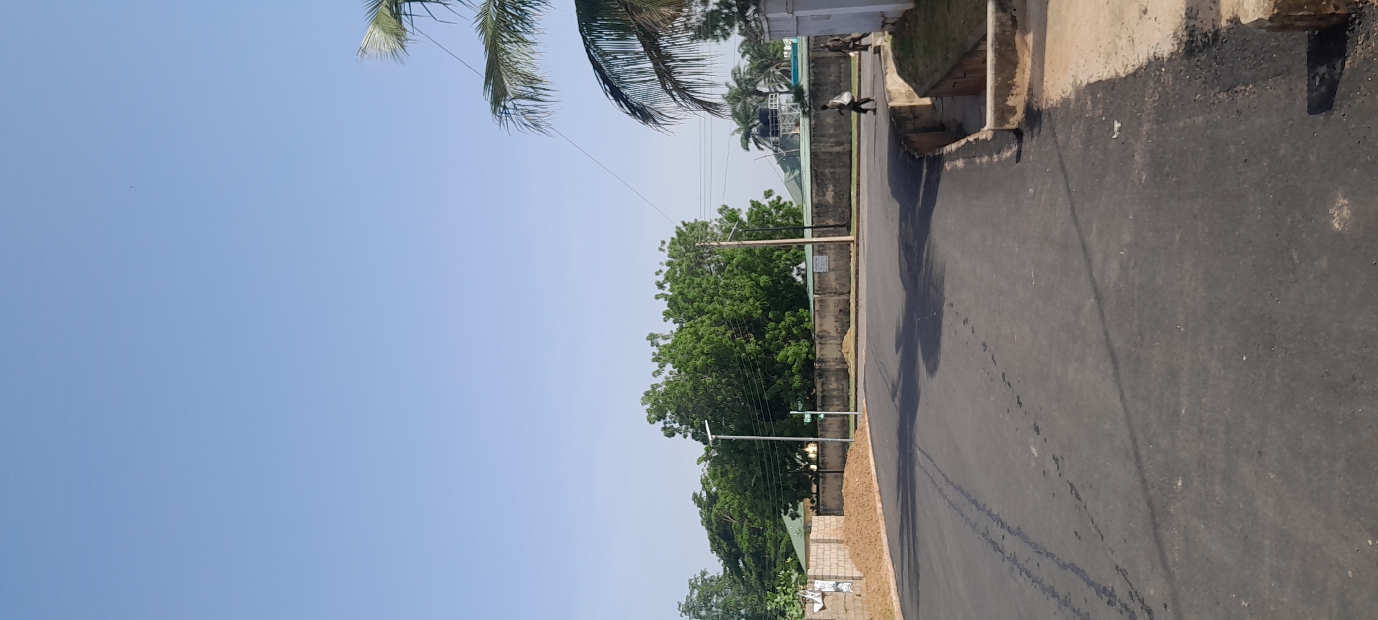 The image shows a paved road curving to the right, likely in Jalingo, Nigeria, under a clear blue sky. On the left side of the road, a pile of sand sits next to a low brick wall. In the background, a long, rough concrete wall runs horizontally, with lush green trees and foliage behind it. A modern street lamp stands tall in front of the wall. Further to the right, a water tank is visible above the wall, and two individuals are walking along the side of the road. One person carries a white bag over their shoulder, while the other walks behind them. Palm trees frame the right side of the image, with their fronds extending into the sky. The road surface is dark asphalt with some lighter-colored streaks and patches.