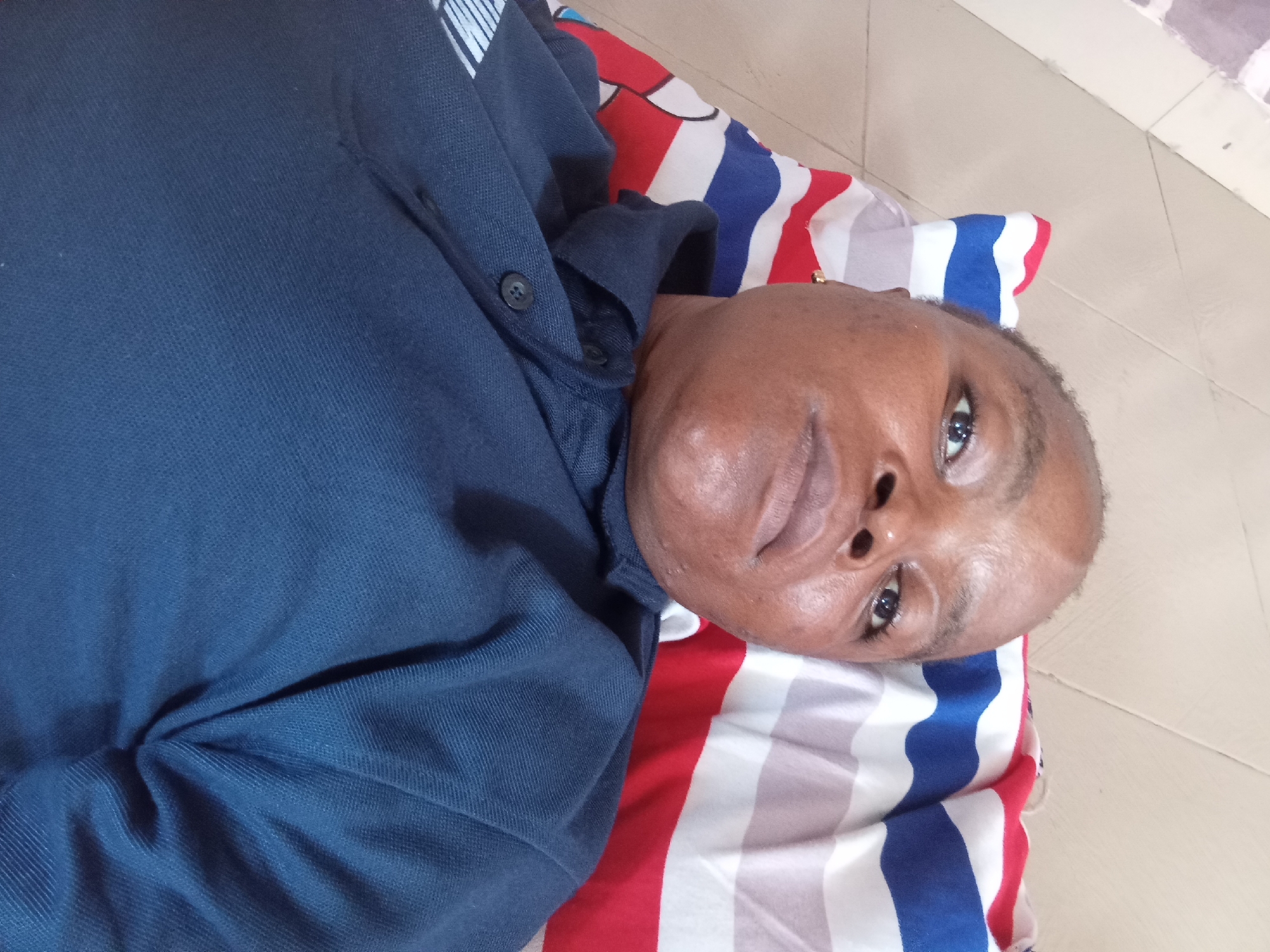 A medium shot captures a Black person lying on their back on a striped pillow. The person is wearing a dark blue collared shirt with two buttons visible on the placket. Their head is turned towards the camera, and they are looking upwards. Their eyes are open, and their mouth is slightly parted. The pillow beneath them is decorated with bold red, white, and blue stripes. The background consists of a tiled floor. The overall impression is that of a person resting or posing for a photograph. The location is Unknown, Unknown.
