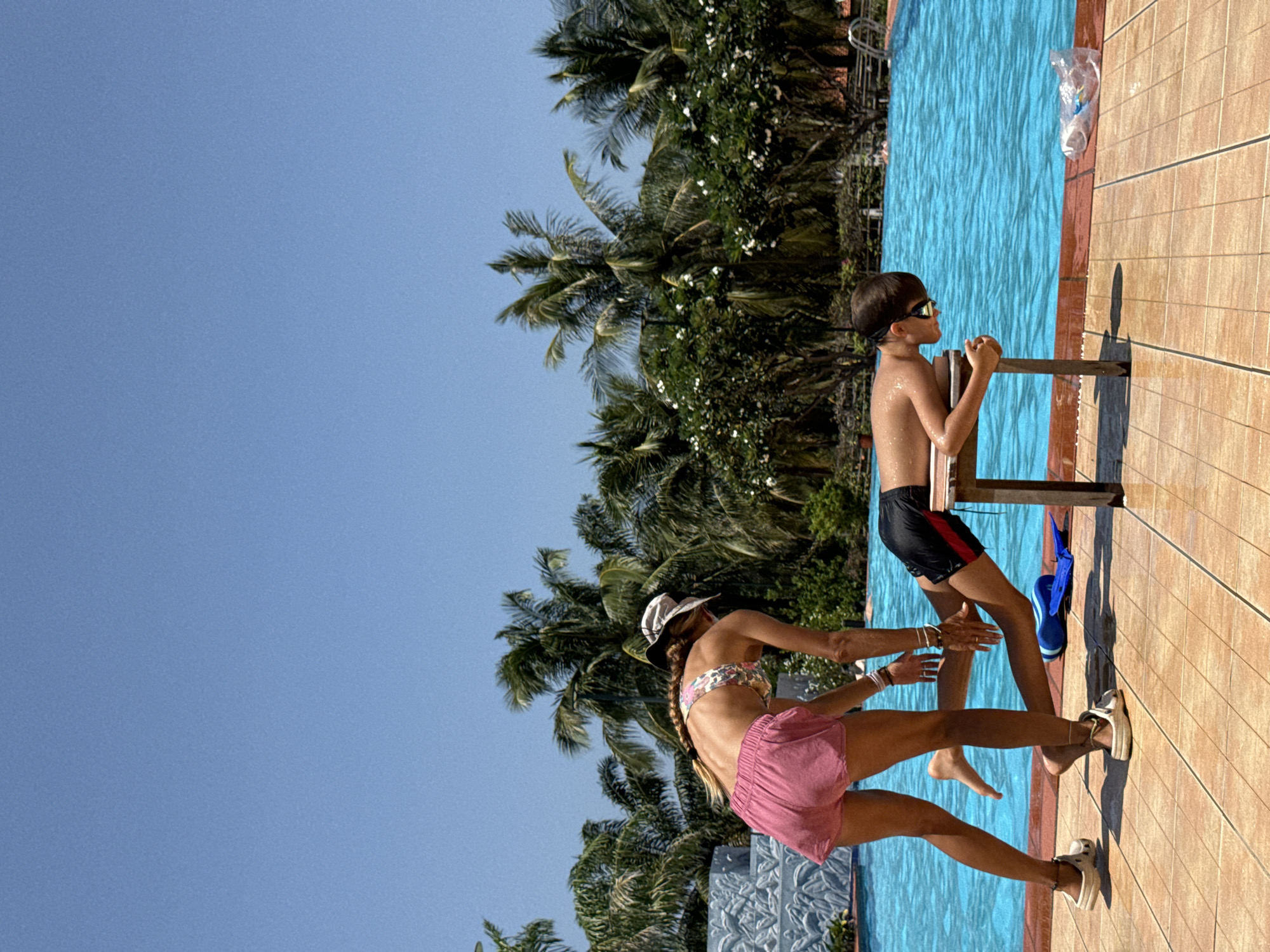 Two children are by a swimming pool on a sunny day. The child on the left, a boy, is wearing swimming goggles and black swim trunks with red trim. He is holding onto a wooden pool ladder, looking out at the water. The child on the right, a girl, is wearing a bikini top with a floral pattern and pink shorts. She has a hat on and her hair in a braid, and she appears to be reaching out towards the boy. They are standing on a tiled pool deck. Behind them, palm trees and lush green foliage are visible against a clear blue sky, suggesting a tropical or subtropical location. The water in the pool is a vibrant blue. A plastic bag is also visible near the edge of the pool. The scene appears to be in the afternoon, given the bright sunlight and shadows.