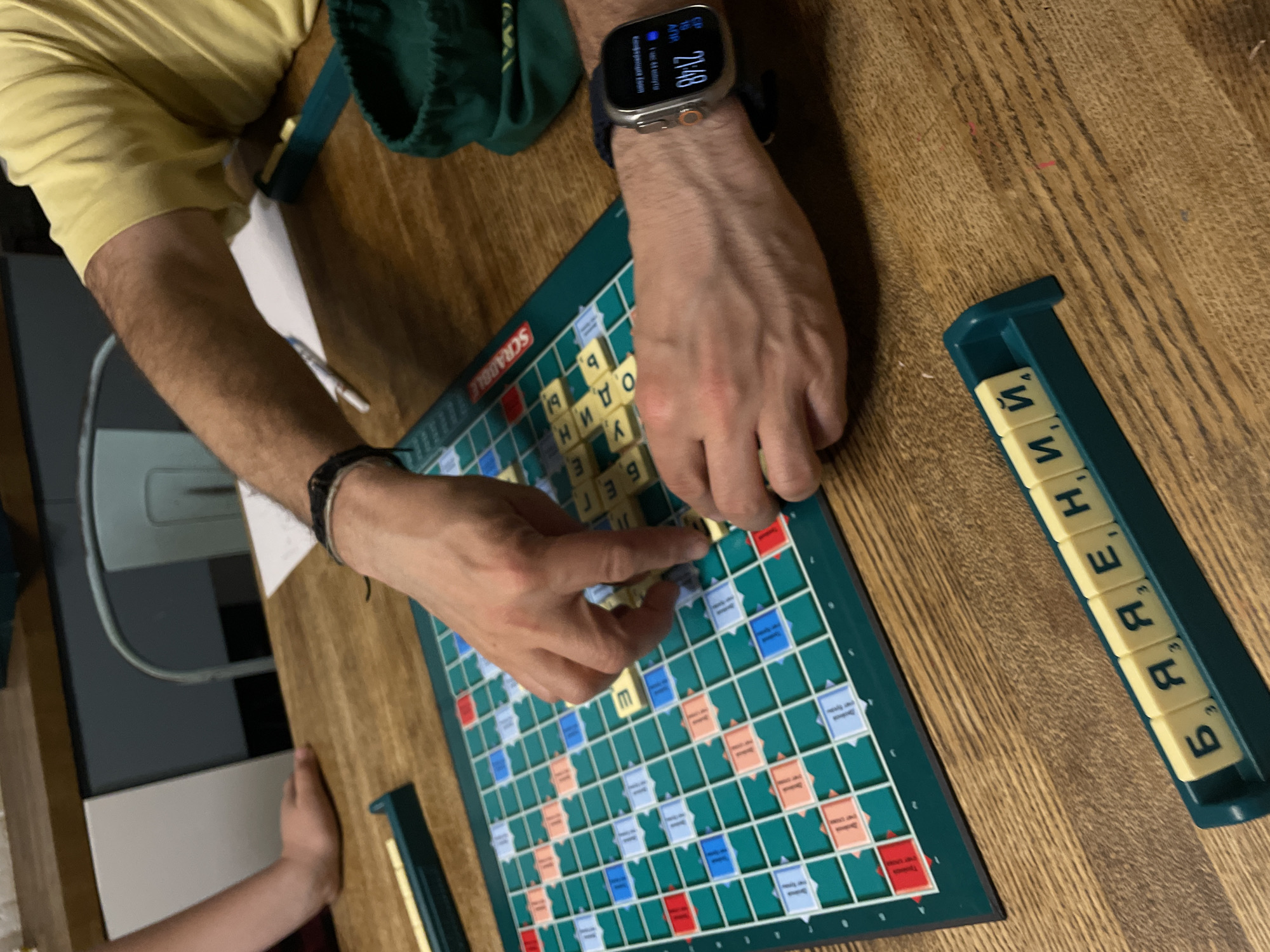 A first-person perspective shot shows two individuals playing the Russian version of Scrabble on a wooden table. One person is actively placing tiles on the game board. The other person's hand is visible near the board. A green bag is present near the first person's left shoulder. The first person wears a yellow shirt and an Apple watch. The setting appears to be indoors. This image does not provide information indicating the location in the city of Moscow, Russia.