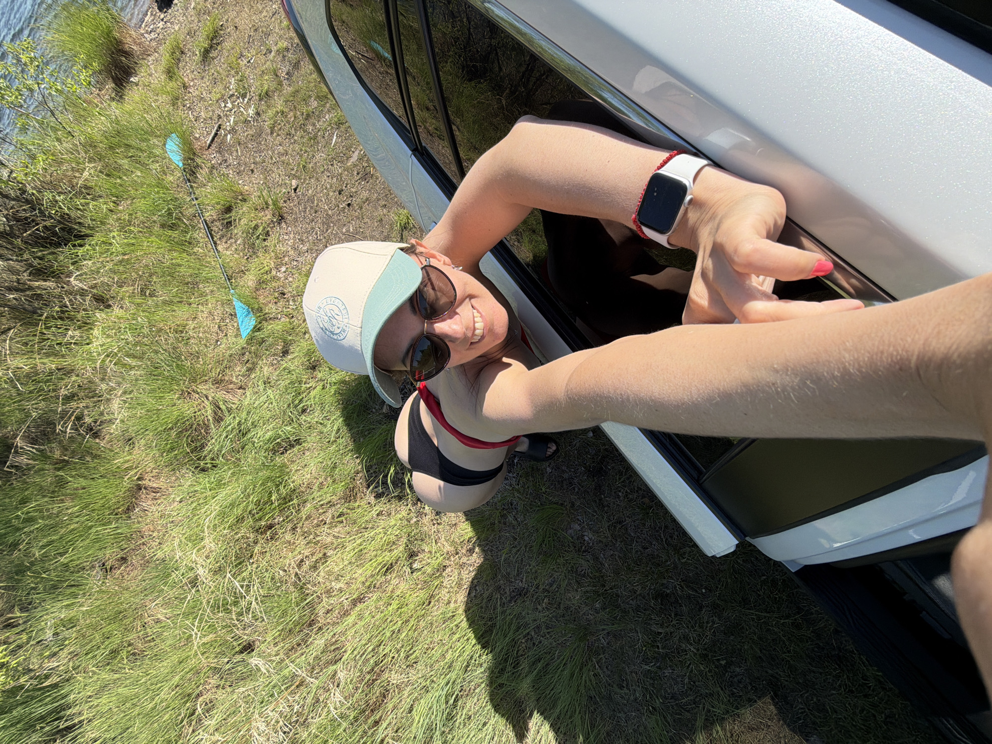 A woman is leaning out of the open door of a white car, smiling at the camera. She is wearing a baseball cap, sunglasses, a swimsuit, and a smart watch. A paddle is laying on the ground near a body of water. The scene takes place in a grassy area near a body of water. The location is not in the city of Birobidzhan, Russia.