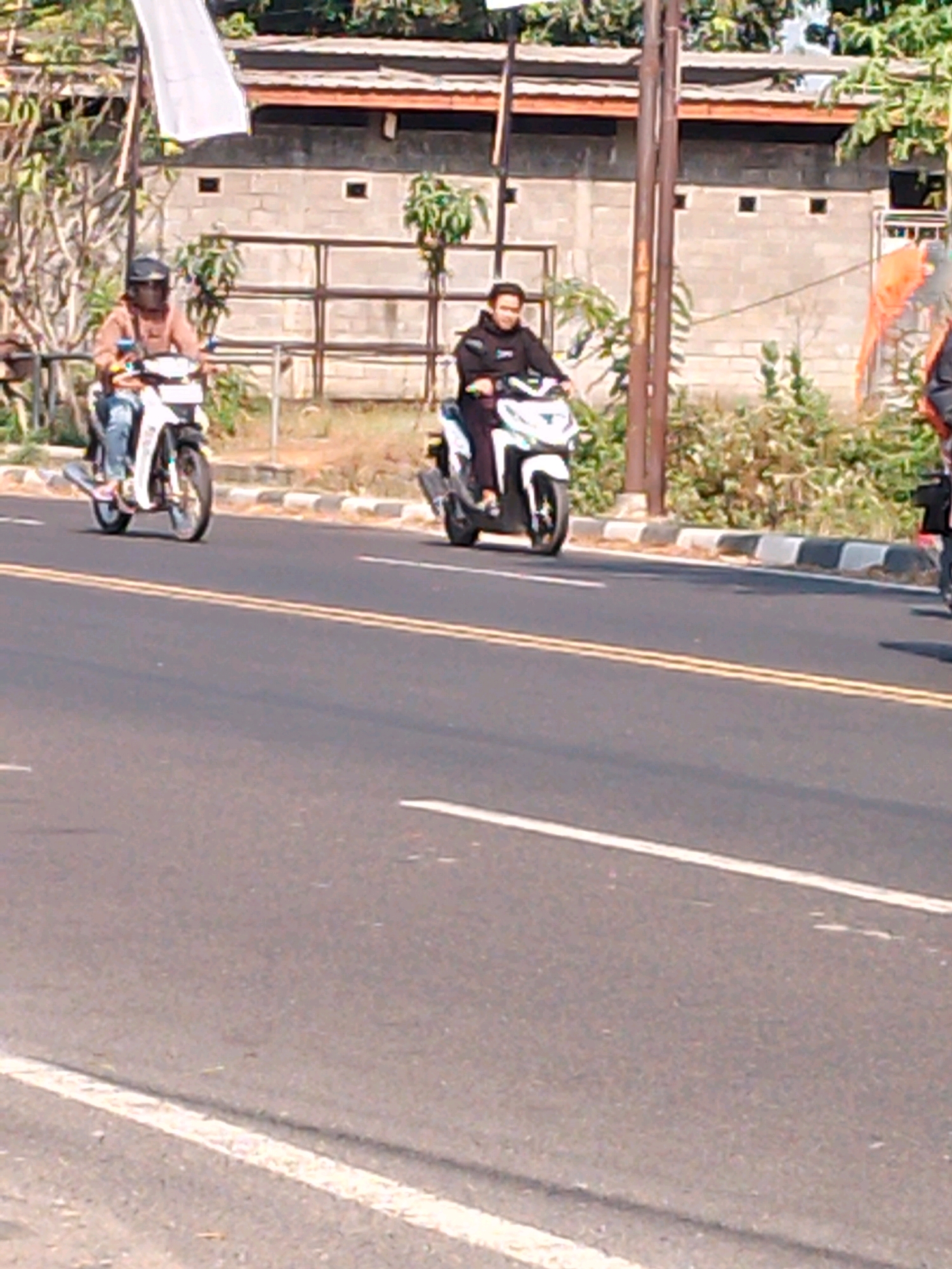 Two individuals on motorcycles are traveling on a paved road in Kutorejo, Indonesia. The person on the left is riding a white motorcycle and is wearing a light brown hooded jacket and a dark helmet with a reflective visor. The person on the right is riding a white scooter and is wearing a dark hooded jacket with a white logo on the left chest. They are on a two-lane road with yellow lines dividing the lanes and white lines marking the edge of the road. In the background, there is a low-rise building constructed of cinder blocks with a reddish-brown roof, and some vegetation. A white flag is visible on the left side of the image. The lighting suggests it is daytime.