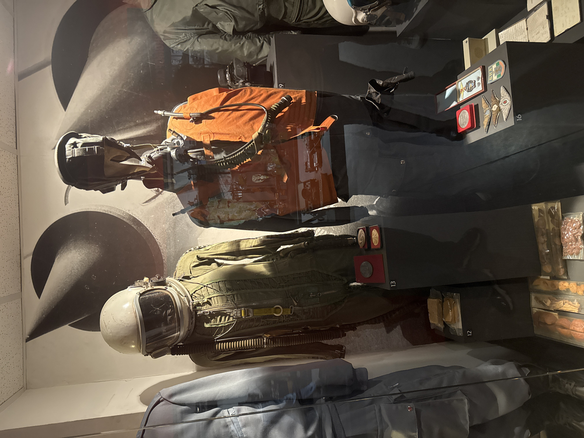 The image captures a display in what appears to be a museum, featuring flight suits and equipment, likely related to aviation or space travel.
The setting seems to be indoors, evidenced by the controlled lighting and what looks like display cases. Two full flight suits are prominently displayed; one is olive green with a rounded helmet and various zippers and attachments, suggestive of a pilot's suit. Below this is a set of blue attire, perhaps regular clothes or another type of uniform. Another flight suit is on display in the upper portion of the image. Behind the flight suits, there is a wall with a large graphic that resembles a portion of a rocket or missile, adding to the overall theme.
On the right side of the display are black panels with information about the objects, including plaques, emblems, or possibly medals associated with the flight suits. Numbers are used to label the objects on display, such as "12", "13", and "16".
Given the Cyrillic writing visible on one of the emblems on the right panel (БНА), it is possible that this is the Bulgarian National Army, and the museum is located in Sofia, Bulgaria.