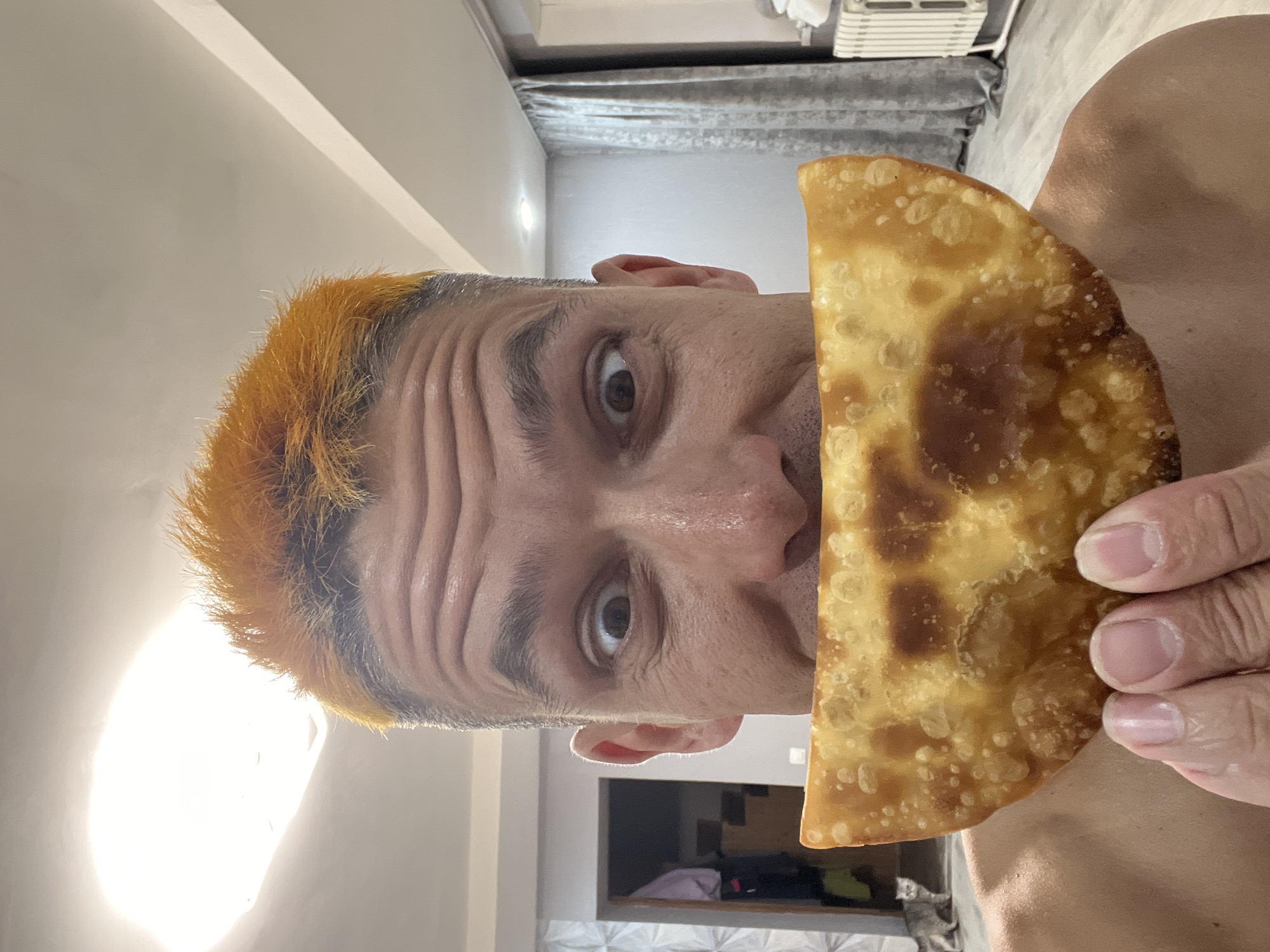 A shirtless man with bright orange dyed hair is holding a fried pastry in front of his face. His eyes are wide and looking directly at the camera. The pastry is golden brown and crescent-shaped, with visible bubbles on its surface. In the background, a doorway reveals a room with some clothes hanging on a rack. To the left of the doorway, a small grey cat is visible, looking towards the camera. The environment appears to be a residential interior, with grey walls, a white ceiling with a light fixture, and a radiator visible on the right. The man is in the foreground, dominating the frame with the pastry obscuring his mouth.