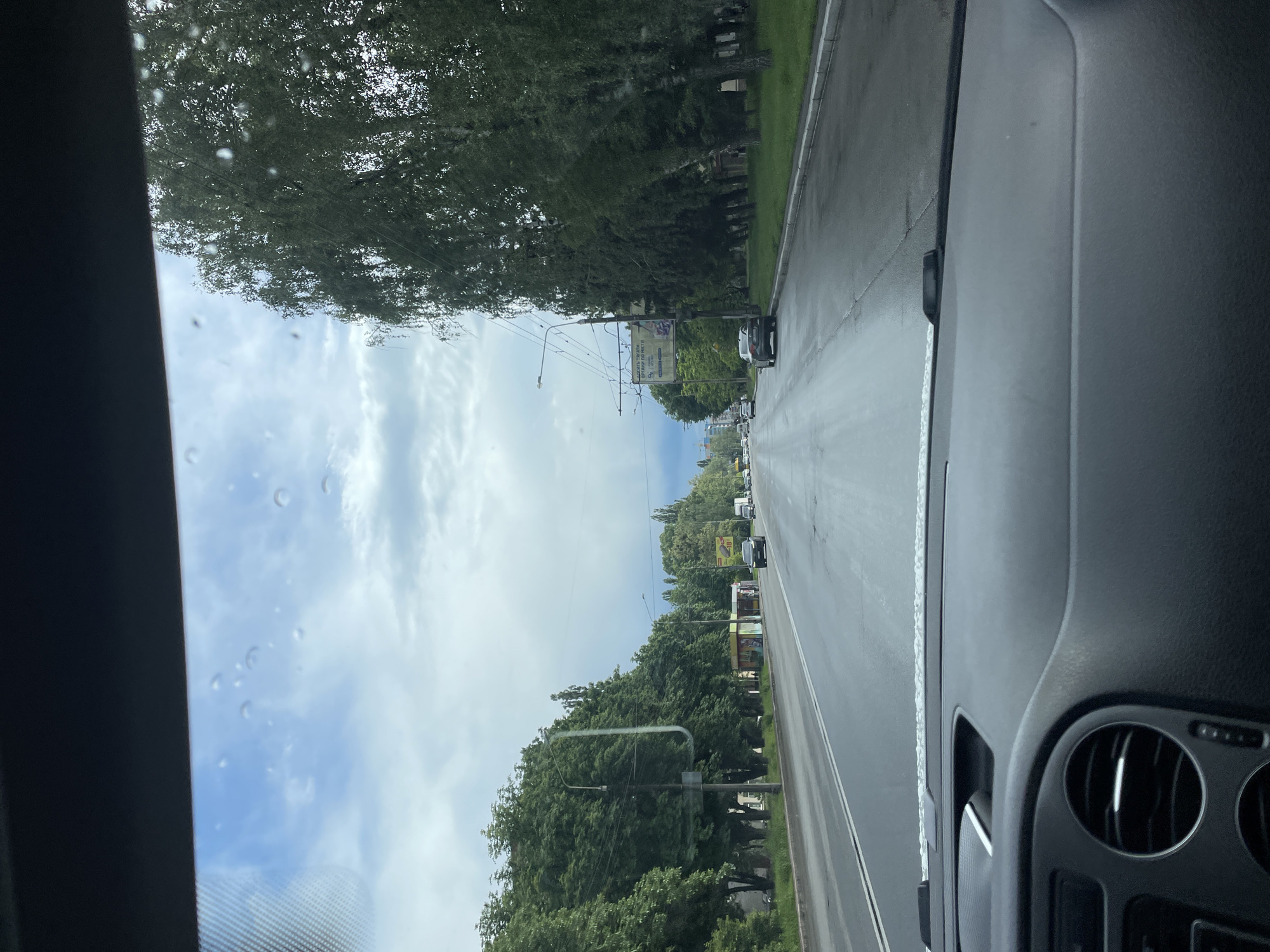 The image shows a street in Poltava, Ukraine, as viewed from inside a car. The street is a long straightaway with multiple lanes of traffic, and cars are visible in the distance. On both sides of the street are rows of trees that line the sides, creating a green canopy. The sky is visible between the trees and is partly cloudy with a mix of blue sky and white clouds. The road surface is wet, likely due to recent rain. The car's dashboard and air vents are visible in the foreground. There are a few water droplets on the windshield. In the background, a street sign or banner can be seen suspended above the road.