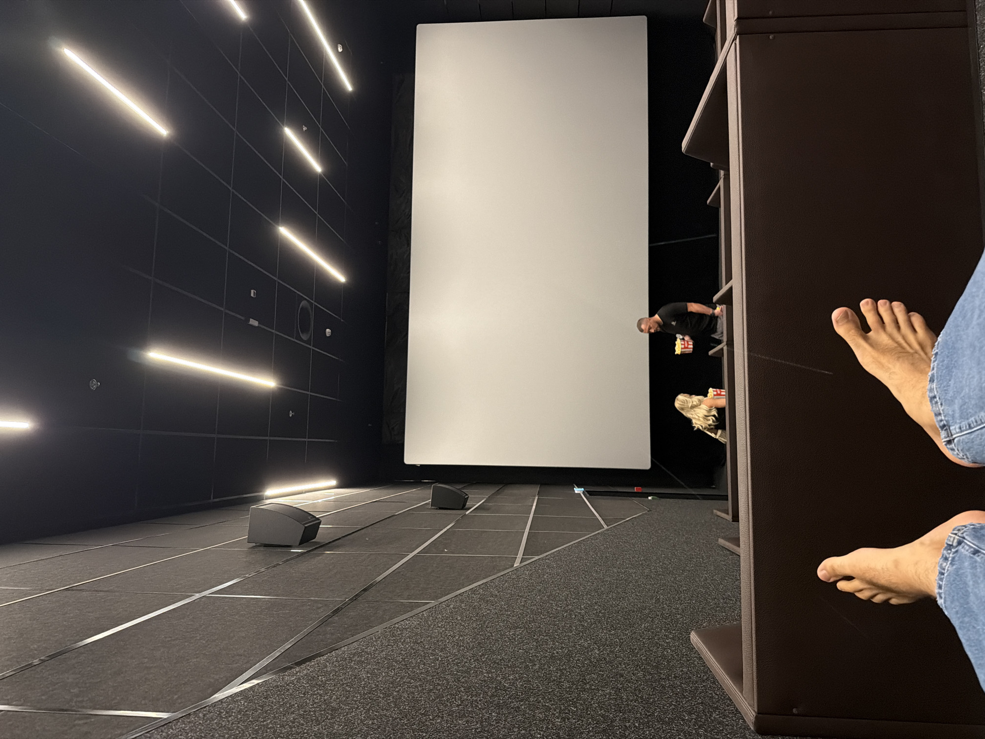 An indoor, low-angle, wide shot shows a movie theater from the perspective of a seated patron. The foreground features the bare feet and lower legs of the photographer, clad in blue jeans. Beyond them, dark brown, upholstered seating for moviegoers is visible. In the mid-ground, a large, blank white movie screen dominates the right side of the frame. To the left of the screen, a blonde woman with her back to the camera is seated and holding a bucket of popcorn. To her right, a man in a black t-shirt and grey shorts is standing and holding a bucket of popcorn. He appears to be interacting with the woman. The walls of the theater are dark grey with a textured surface and diagonal silver metallic strips. The ceiling is black with a grid pattern and illuminated by long, linear fluorescent lights. The scene is dimly lit, consistent with a movie theater environment. The photographer's location is a movie theater in the city of Kyiv, Ukraine.