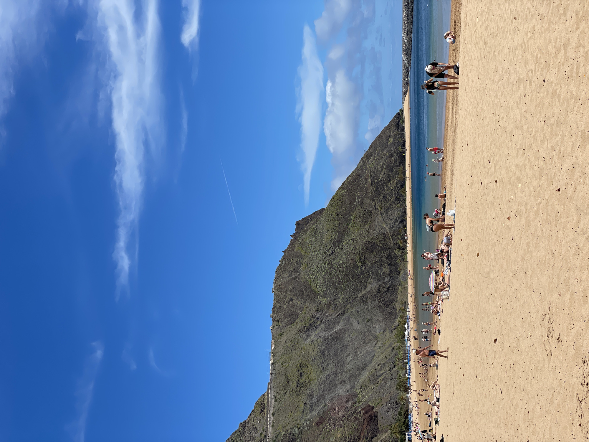 This image depicts a sunny and vibrant beach scene at El Suculum, Spain, likely during the daytime under clear weather conditions. The sky is a bright, deep blue with scattered, elongated white clouds, and a subtle airplane contrail is visible in the upper portion.
To the left, a large, steep mountain or hill rises dramatically, its rugged slopes covered in dark green vegetation and rock formations. A faint line, possibly a road or trail, can be seen along its base, with some small structures dotted further up.
The right side and foreground are dominated by a wide expanse of golden-brown sand, densely populated with numerous people. Individuals are engaged in various beach activities, including sunbathing, walking, and relaxing. Some are sitting or lying on towels, while others are gathered around beach umbrellas.
The calm, clear turquoise waters of the sea gently lap against the shore. Many people are visible in the ocean, swimming, wading, or standing in the shallows, with more figures scattered further out. The overall impression is one of a busy and enjoyable day at a popular coastal destination. No visible text is present in the image.
