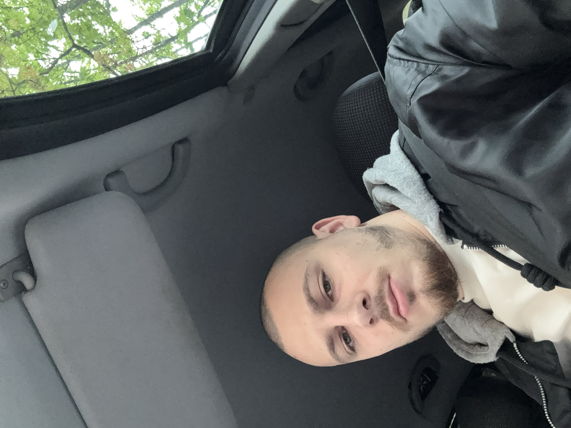 A selfie captures a man from inside a car in Stara Zagora, Bulgaria. The man, positioned in the lower right portion of the frame, has a shaved head, a light beard, and is looking towards the camera with a neutral expression. He is wearing a grey hooded sweatshirt underneath a black jacket.
The setting is the interior of a car, seen from an upward angle. Features of the car include a light grey headliner, a sun visor on the left, and grey grab handles. Part of a dark grey, mesh-patterned car seat is visible behind the man. The car is equipped with a sunroof, through which green tree foliage and branches are visible, suggesting it is daytime and the car is outdoors. No specific activity beyond taking the selfie is depicted. No text is visible in the image.