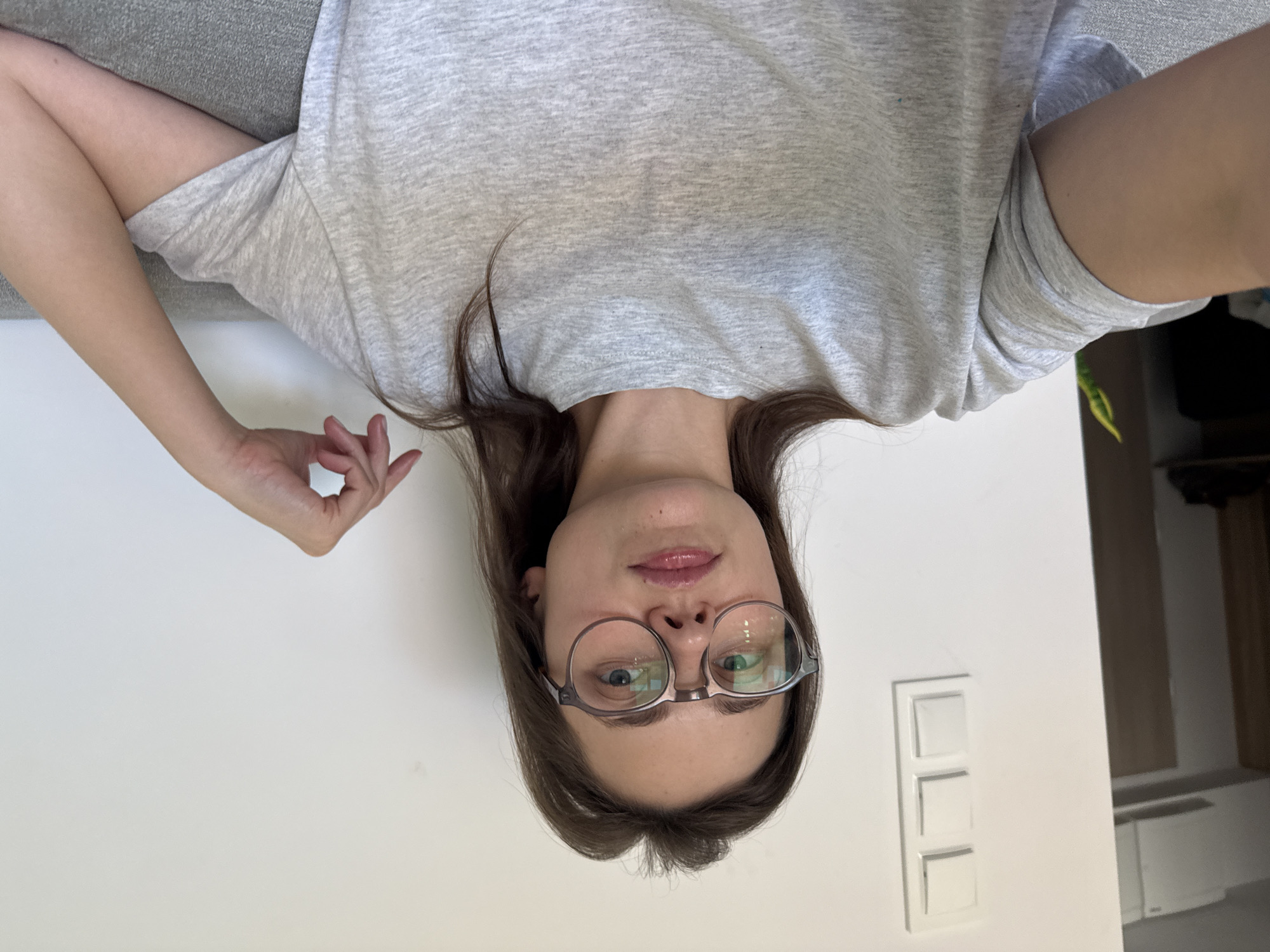 The image shows a woman wearing glasses and a grey t-shirt. Her head is tilted to the side, and her hair is dark brown and falls around her face. The woman is looking at the camera, and her mouth is slightly open. The background is a plain white wall with a light switch. The image appears to be taken indoors, possibly in a home or apartment. The lighting suggests it is daytime. There is no visible text in the image. The overall impression is a casual, candid portrait.