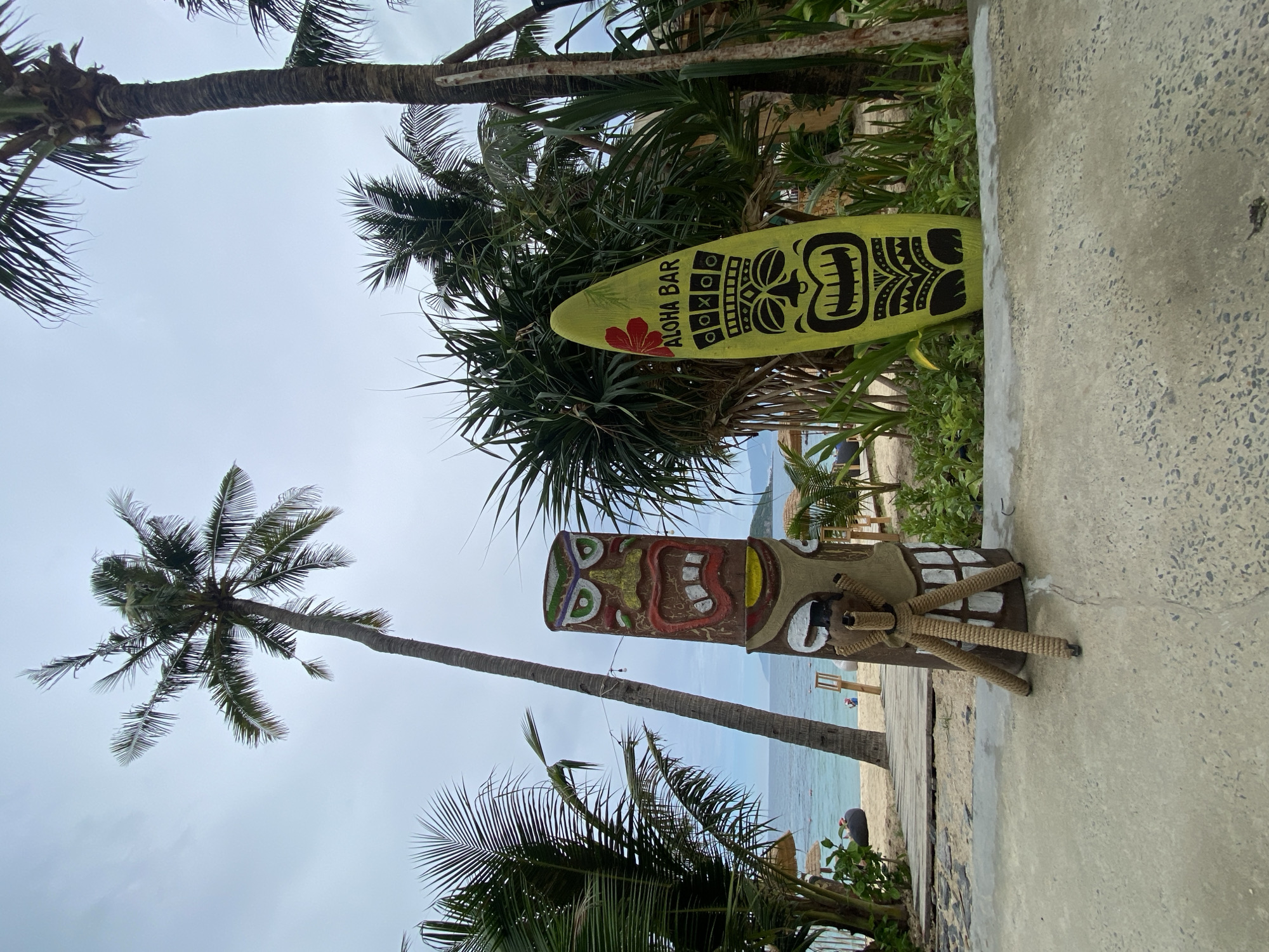 This image depicts a tropical beach scene, likely in Nha Trang, Vietnam, given the presence of tiki-style decor and palm trees. In the foreground, a carved wooden tiki statue stands on a concrete pathway, with a tripod-like structure made of rope at its base. To the right of the tiki statue, a bright yellow surfboard with a traditional tiki face design and the text "ALOHA BAR" adorned with a red flower leans against lush green foliage. In the background, a sandy beach stretches towards the ocean, with a wooden walkway leading to the water. Several palm trees are visible, including a tall one in the center of the frame. The sky is overcast. The ocean is calm with a few distant boats. There are some thatched umbrellas and seating visible on the beach, as well as a few people in the distance. The overall atmosphere is relaxed and vacation-like.