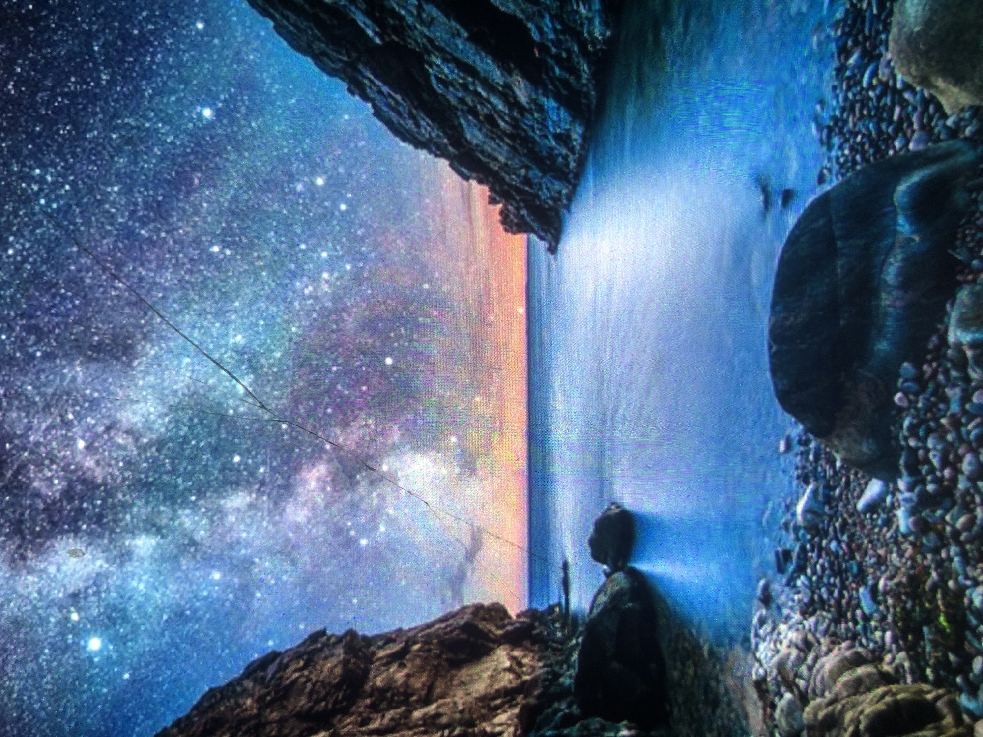 The image depicts a surreal and dreamlike landscape, not a scene from Tashkent, Uzbekistan. It features a body of water nestled between two imposing, rocky cliffs. Above, a vibrant night sky is ablaze with a dense array of stars, transitioning into a colorful twilight horizon with hues of orange and pink. The water reflects the celestial display and the surrounding cliffs, creating a sense of depth and wonder. The foreground is a rocky shoreline, with various sizes of stones and pebbles scattered along the edge of the water. The overall impression is one of natural grandeur and cosmic beauty, evoking a sense of peace and awe.