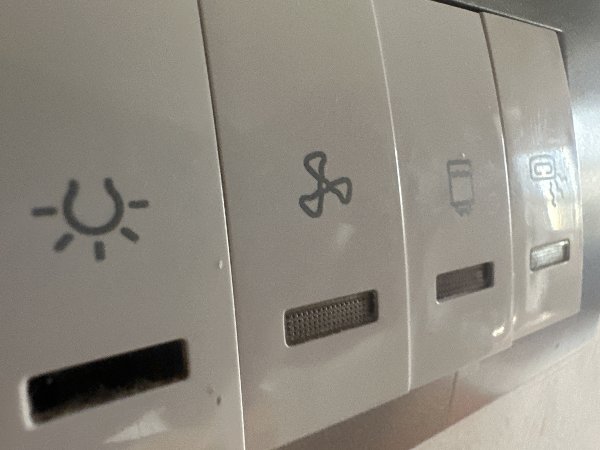 This is a close-up, eye-level shot of a section of a light-colored, plastic control panel, likely found in a vehicle or an appliance. The panel has several distinct icons and features etched or printed onto its surface, suggesting different functions.
From left to right, the visible elements are:
1. **Light Bulb Icon:** A stylized representation of a light bulb with radiating lines, indicating a lighting function. Below this icon, there is a dark, rectangular slot, possibly for a USB port or a card reader.
2. **Fan Icon:** A three-bladed fan symbol is centered in the next section. Beneath this icon is a textured, rectangular opening, likely a vent or speaker grille.
3. **Battery Icon:** The third section displays an icon resembling a battery, with a plus and minus sign, suggesting a power-related function or battery status indicator.
4. **C/Charging Icon:** The furthest visible section shows an icon that appears to be a "C" inside a rounded square, with a wavy line underneath, commonly representing a charging port or a charging indicator. There is also a small, horizontal slot above this icon, similar to the one seen with the light bulb icon.
The control panel is clean overall, with some minor scuffs and dust visible, particularly on the left side. The lighting in the image is bright, suggesting it was taken in a well-lit environment, possibly indoors. The overall aesthetic is utilitarian and modern. No people are visible in the frame. The context of Belgrade, Serbia is noted but not visually represented in the image itself.