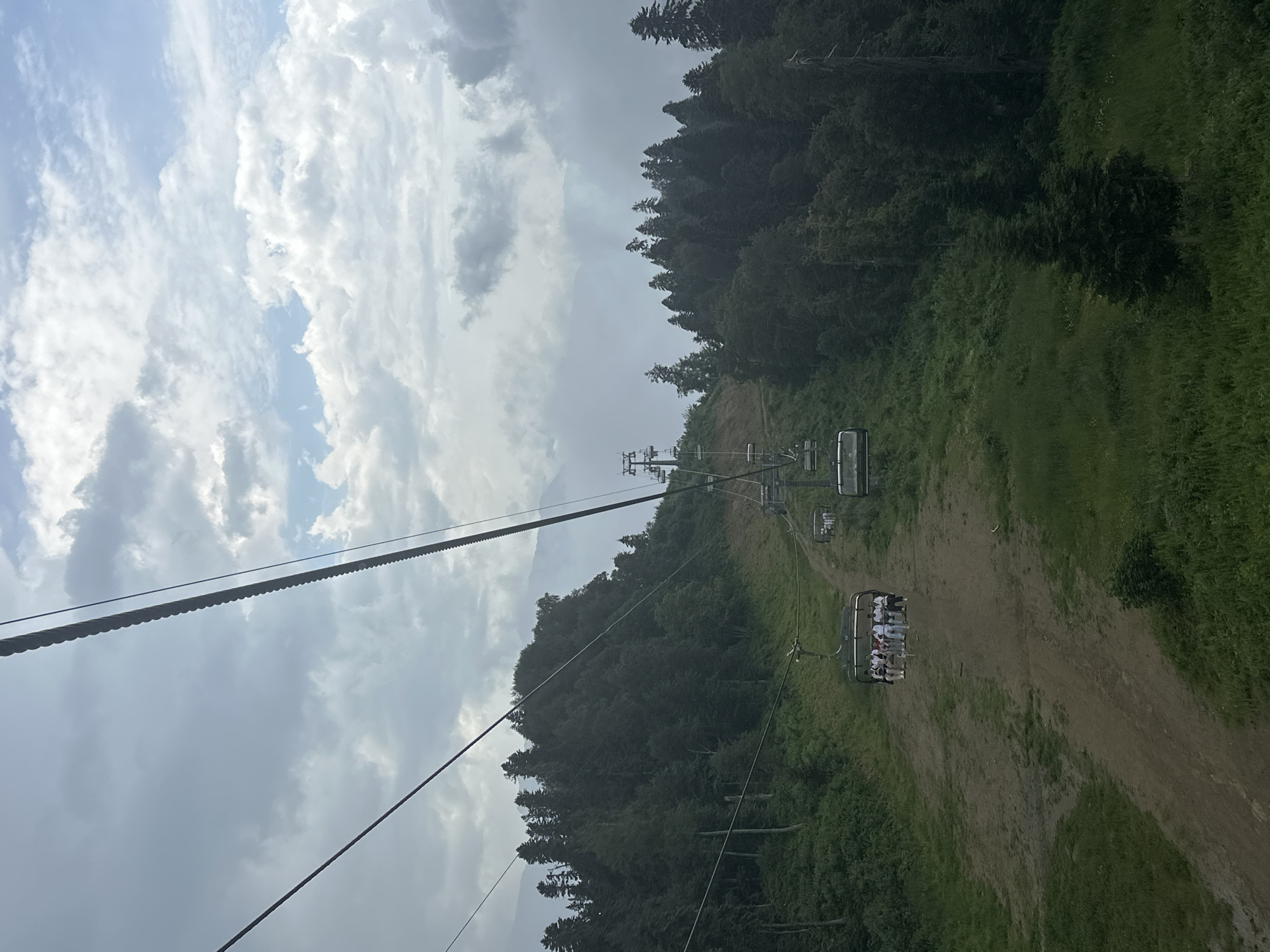 The image depicts a ski lift ascending a mountainous, tree-covered slope in Estosadok, Russia. The sky is overcast with large, white clouds. In the foreground, a ski lift cable stretches diagonally across the frame. Several empty ski lift chairs are visible on the cable, along with one chair carrying a group of people. The slope is covered in green grass and a dirt path. In the mid-ground, dense green trees cover the hillside. The background shows more tree-covered mountains shrouded in mist. The overall impression is of a cloudy, cool day at a mountain resort.
