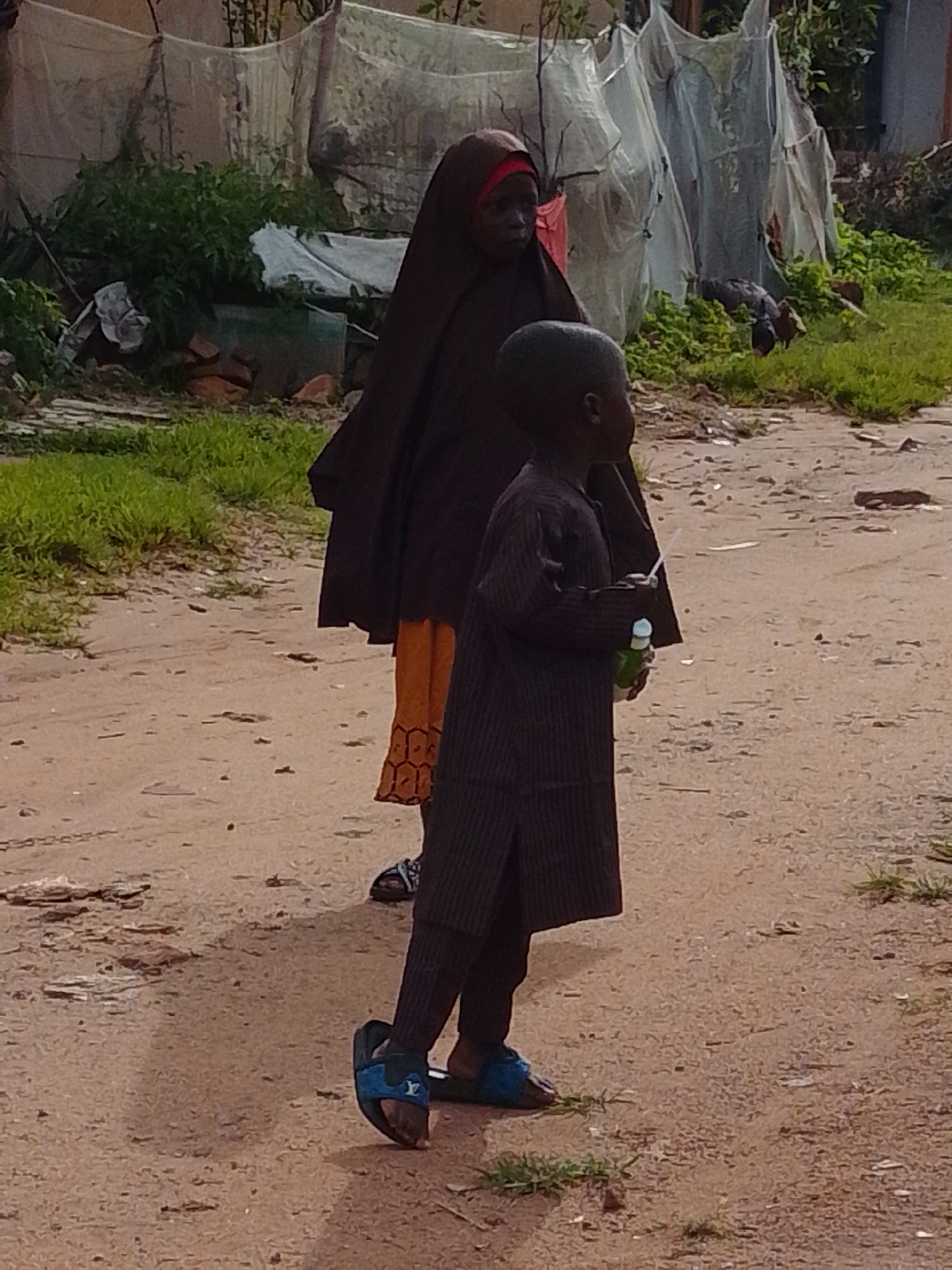 A medium shot captures two figures in a dusty, outdoor environment, likely in Gombe, Nigeria. In the foreground and slightly to the right, a young boy stands with his back to the camera, looking to the left. He is wearing a dark, pinstriped tunic and blue slide sandals. In his right hand, he holds a green bottle with a white straw. Behind and to the left of the boy stands a woman wearing a dark brown hijab that covers her head and body, with a glimpse of an orange garment visible beneath. She is also looking to the left. The ground is a mix of sandy dirt and sparse green grass. In the background, there are structures covered with white netting, suggesting possibly a market stall or agricultural netting, with some greenery and scattered debris.