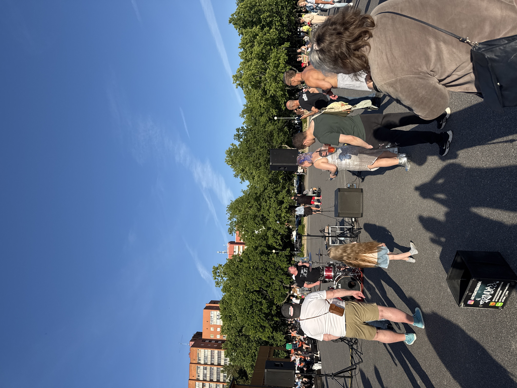 The image depicts an outdoor gathering with a band performing on a stage in the city of Tallinn, Estonia. The sky is clear and blue, with scattered white clouds. Tall, leafy green trees form a backdrop behind the stage, and several multi-story brick buildings are visible in the distance.
In the foreground, a group of people are gathered, some standing and watching the performance, while others are seated on the ground. A band is set up on a makeshift stage, with drums, microphones, and speakers visible. A male vocalist is singing into a microphone, and other band members are implied but not clearly visible.
Several individuals are the focus of attention. A man in a white t-shirt and khaki shorts stands with his back to the camera, looking towards the performance. To his left, a young girl with long blonde hair wears a denim skirt and t-shirt. Several other attendees are scattered throughout the scene, some holding drinks, and others engaging in conversation. The ground is paved asphalt, and long shadows are cast by the people and objects, indicating it is late afternoon or early evening. A black bin with a sticker advertising "ARTEMTEMADA" is in the lower left foreground.