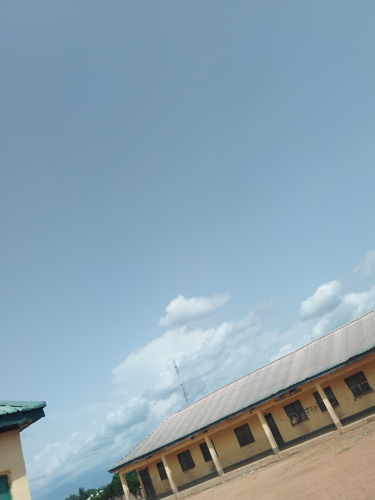 The image shows a view of a school building in Serti, Nigeria. The building is a single-story structure with a corrugated metal roof and yellow walls. It has several windows and doors. The school's name, "BASHAKA L.G.C. 2008", is visible on the side of the building. The sky is a clear blue with some white clouds. There is also a small building with a green roof visible in the foreground on the left. A tall radio tower can be seen in the background on the right. The environment appears to be somewhat rural, with some trees and vegetation visible in the distance. There are no people visible in the image. The overall impression is of a sunny day at a school.