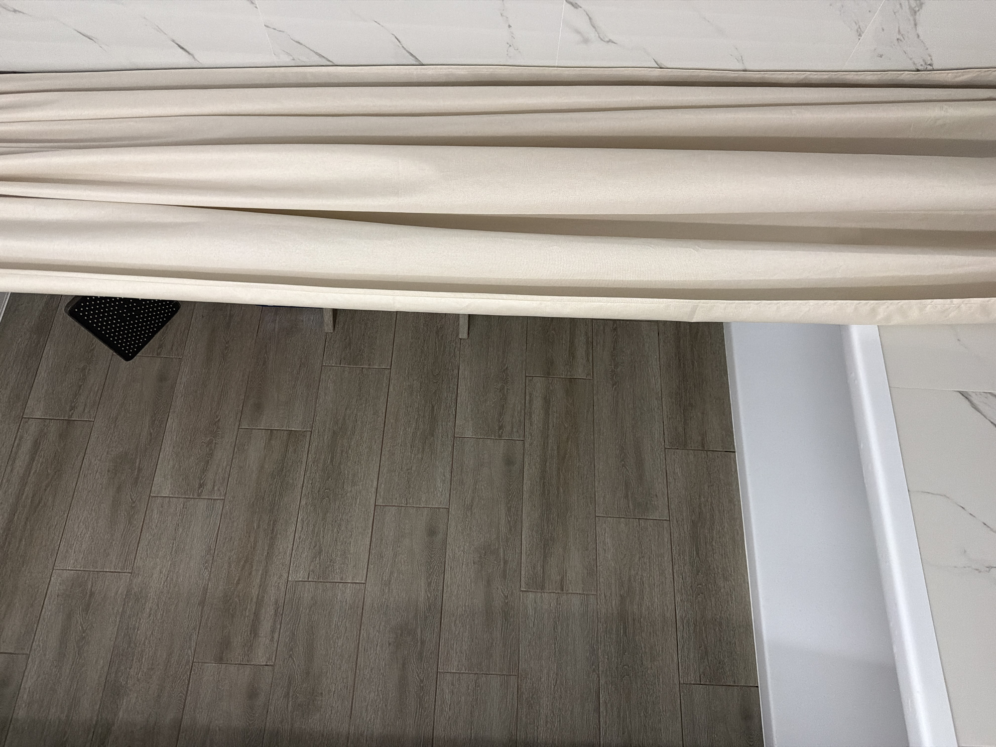 The image depicts a bathroom scene. The floor is covered in wood-patterned tiles, and a black rubber bath mat is visible near a white bathtub. The tub is adjacent to a wall tiled with white marble. A beige shower curtain obscures part of the view.
It is not possible to determine the location of the bathroom in Chaikovsky, Russia from the image.