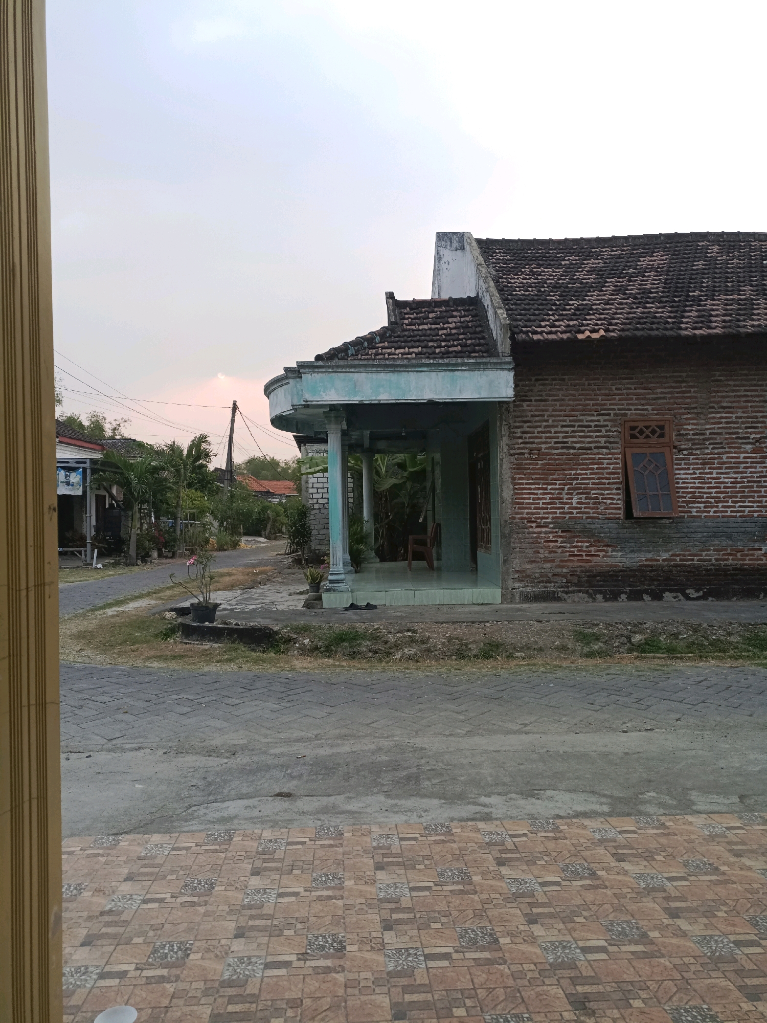 The image depicts a street scene in Rancang Kencono, Indonesia, during a late afternoon or early evening. The sky is a pale, hazy pink, suggesting a sunset or sunrise with some atmospheric obstruction. In the foreground, a tiled walkway leads towards a paved road. To the right, a brick house with a dark tile roof is visible. The house features a covered porch supported by light blue columns. A wooden window with a grid pattern is set into the brick wall of the house.
The road curves into the distance, lined with various houses and vegetation, including palm trees and other tropical plants. Utility poles with wires are present along the road. On the left side of the road, another house is partially visible, with a blue sign attached to its exterior. A small potted plant sits on a low concrete structure near the road. The overall environment suggests a residential area in a warm climate, possibly a village or a less developed part of a town. No people are visible in the image.