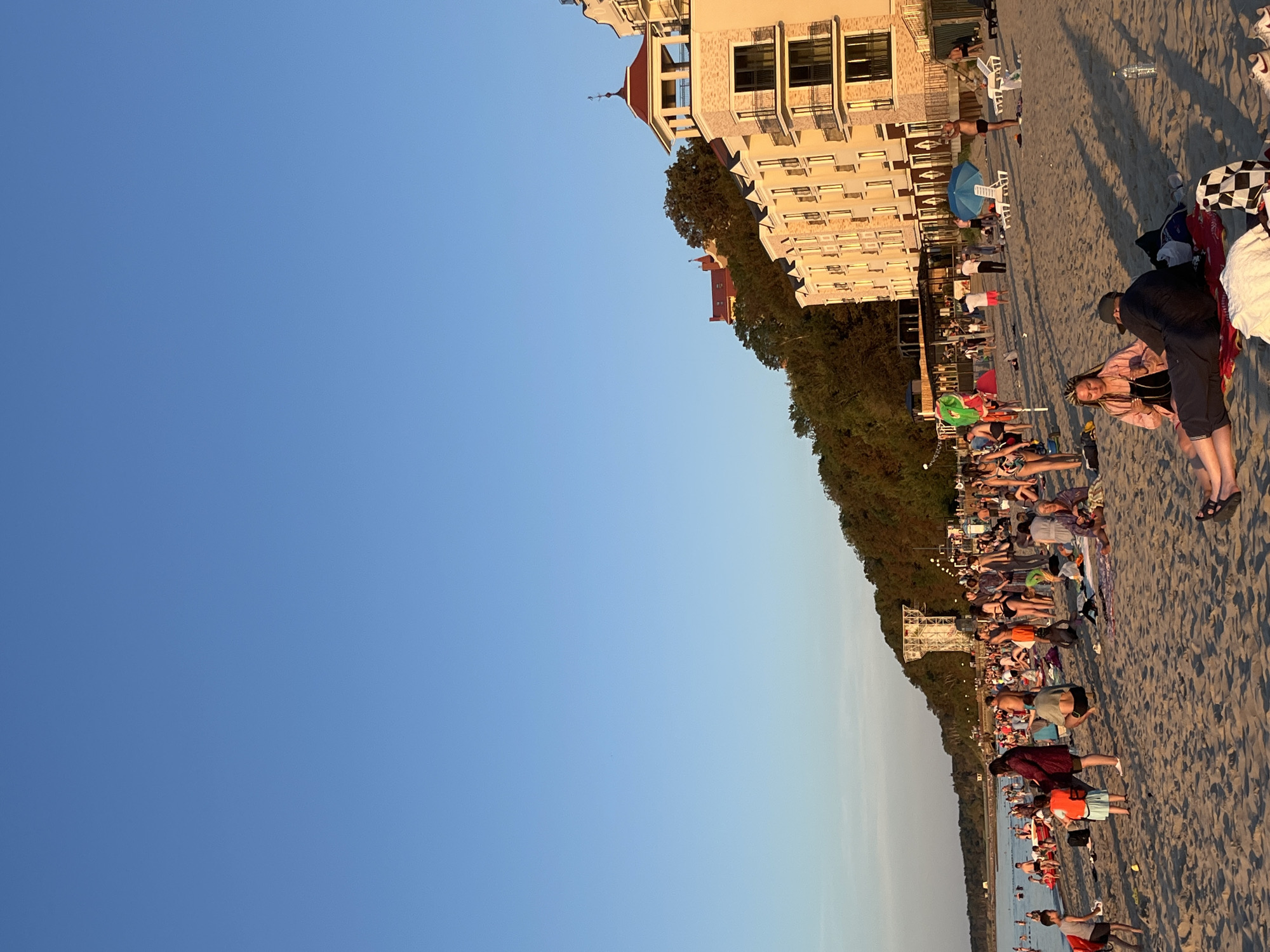 The image depicts a crowded beach in Pionersky, Russia, under a clear blue sky. The sandy beach is populated with numerous people engaged in various activities. Some are sunbathing on towels and loungers, while others are sitting or standing, socializing. Children are visible playing on the sand. In the background, the sea stretches out, and beyond the beach, a lush green hillside is topped with several buildings, including a distinct light-colored hotel with multiple balconies and a red-domed tower. The setting appears to be a warm, sunny day, with long shadows cast across the sand, suggesting it is late afternoon or early evening.