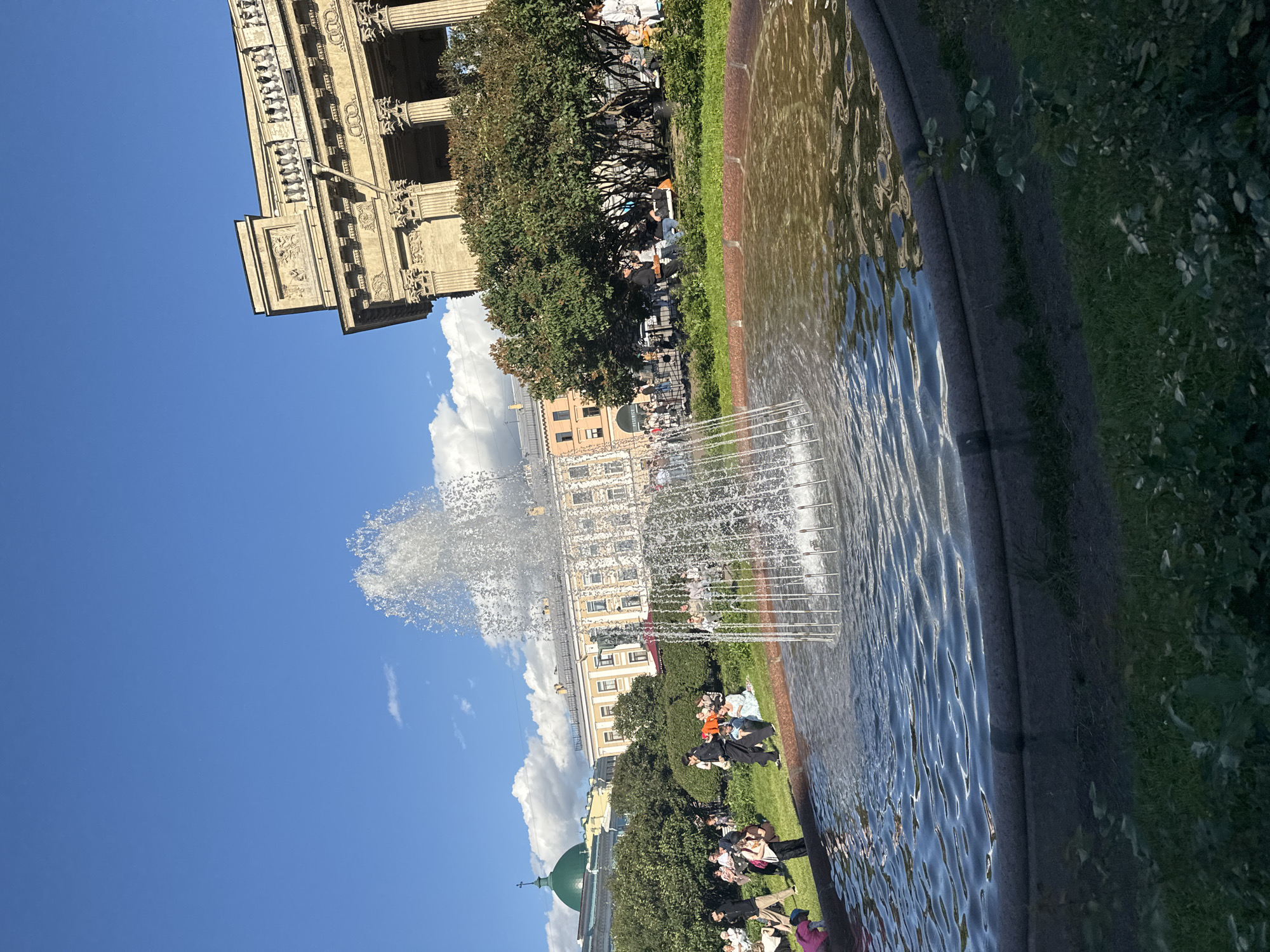 The image depicts a sunny day in a public square in Saint Petersburg, Russia, likely Kazan Cathedral's colonnade. In the foreground, a circular fountain with a spouting water jet is prominently featured, its water reflecting the blue sky and surrounding greenery. The fountain basin has a dark gray stone rim and is surrounded by green grass.
In the middle ground, a diverse group of people are gathered in the grassy area, some sitting, others standing, and a few walking. The attire of the individuals suggests a casual, leisurely atmosphere. Behind the people and the fountain, lush green trees form a dense line.
The background showcases a grand, neoclassical building with a beige facade, adorned with columns and ornate architectural details, consistent with the style of Kazan Cathedral. To the left of this building, a green domed structure, possibly a church or another part of the cathedral complex, is visible. The sky is a vibrant blue with scattered white clouds, indicating clear weather. The overall scene conveys a sense of urban recreation and historical architecture.