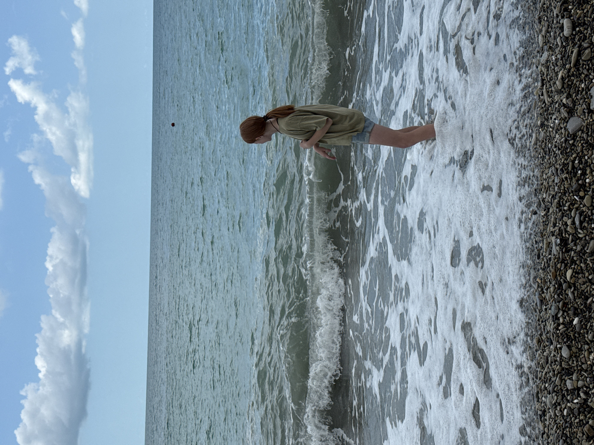 In Nebug, Russia, a young woman with reddish-brown hair, tied back in a ponytail, stands in the shallow surf on a pebbled beach. She is wearing a light green t-shirt and denim shorts. The water is a cool blue-green with moderate waves, and white foam washes over her legs. The sky above is a pale blue, dotted with fluffy white clouds. A small red buoy is visible in the distance on the water. The foreground is dominated by the texture of the pebbles on the beach.