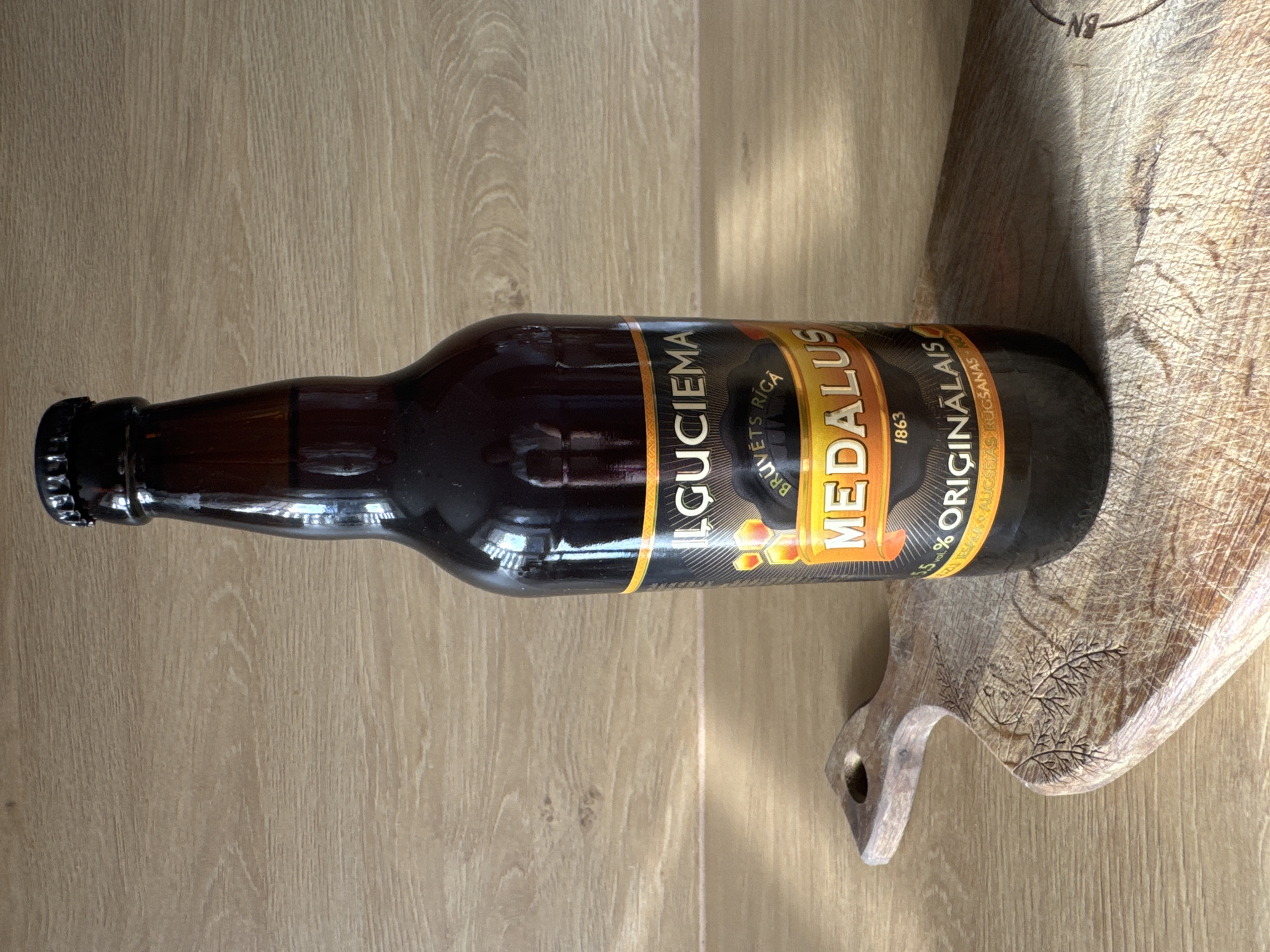 A dark brown glass beer bottle with a black cap stands upright on a rustic wooden cutting board. The cutting board has a carved design of leaves and is placed on a light-colored wooden surface. The bottle's label features text in gold and black, including "ILGUIECIEMA", "MEDALUS", and "ORIGINĀLAIS", along with a year "1863". The scene is lit from the left, casting a shadow from the bottle onto the cutting board and the surface behind it. The background is a plain, light-colored wooden wall. The overall impression is a still life arrangement, possibly in a kitchen or dining area. There are no people visible in the image. The scene appears to be indoors, with diffused natural light creating highlights and shadows. The city of Riga, Latvia is not depicted or suggested by the visual elements in this image.