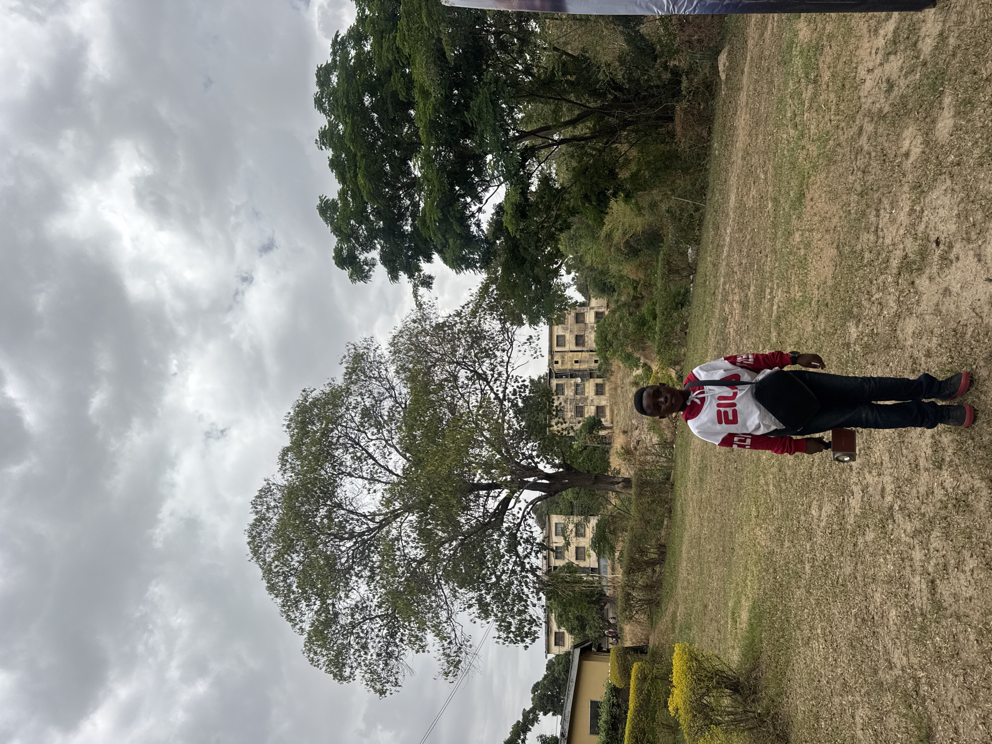The image shows a person standing on a grassy field in Jos, Nigeria. The person is wearing a white sweatshirt with red sleeves and black pants. The sky is overcast. There are buildings in the background behind the tree on the left, and there are some trees in the background. A yellow building and bushes are in the foreground. The ground appears dry and the vegetation is parched. The person is holding an object in their right hand.